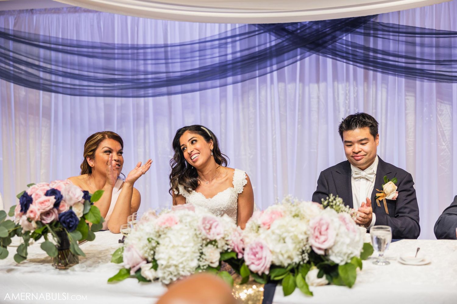 Wedding party seated at head table with pink and white floral centerpieces and blue draping.