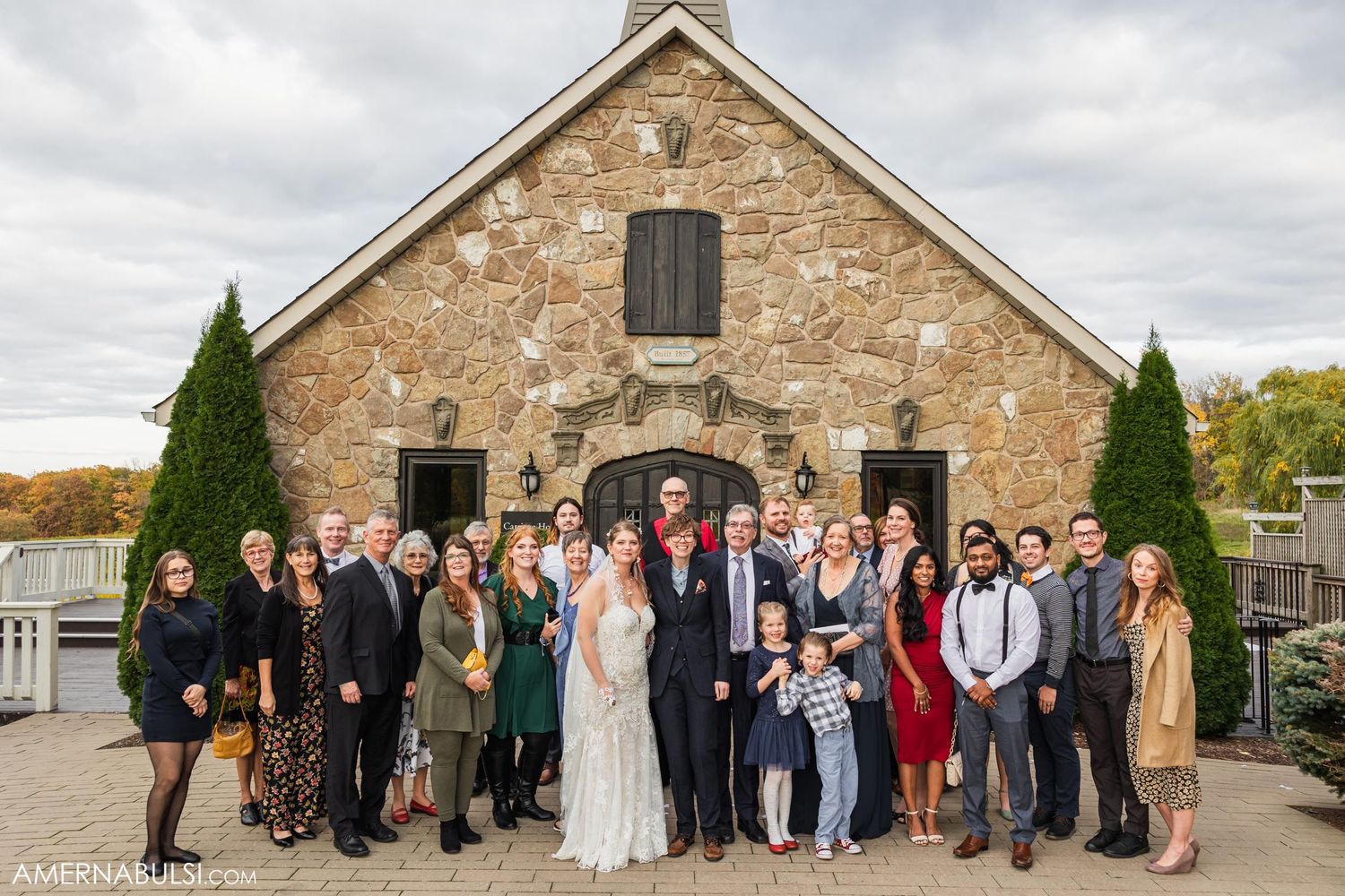Wedding party group photo in front of rustic stone barn with fall colors in background.