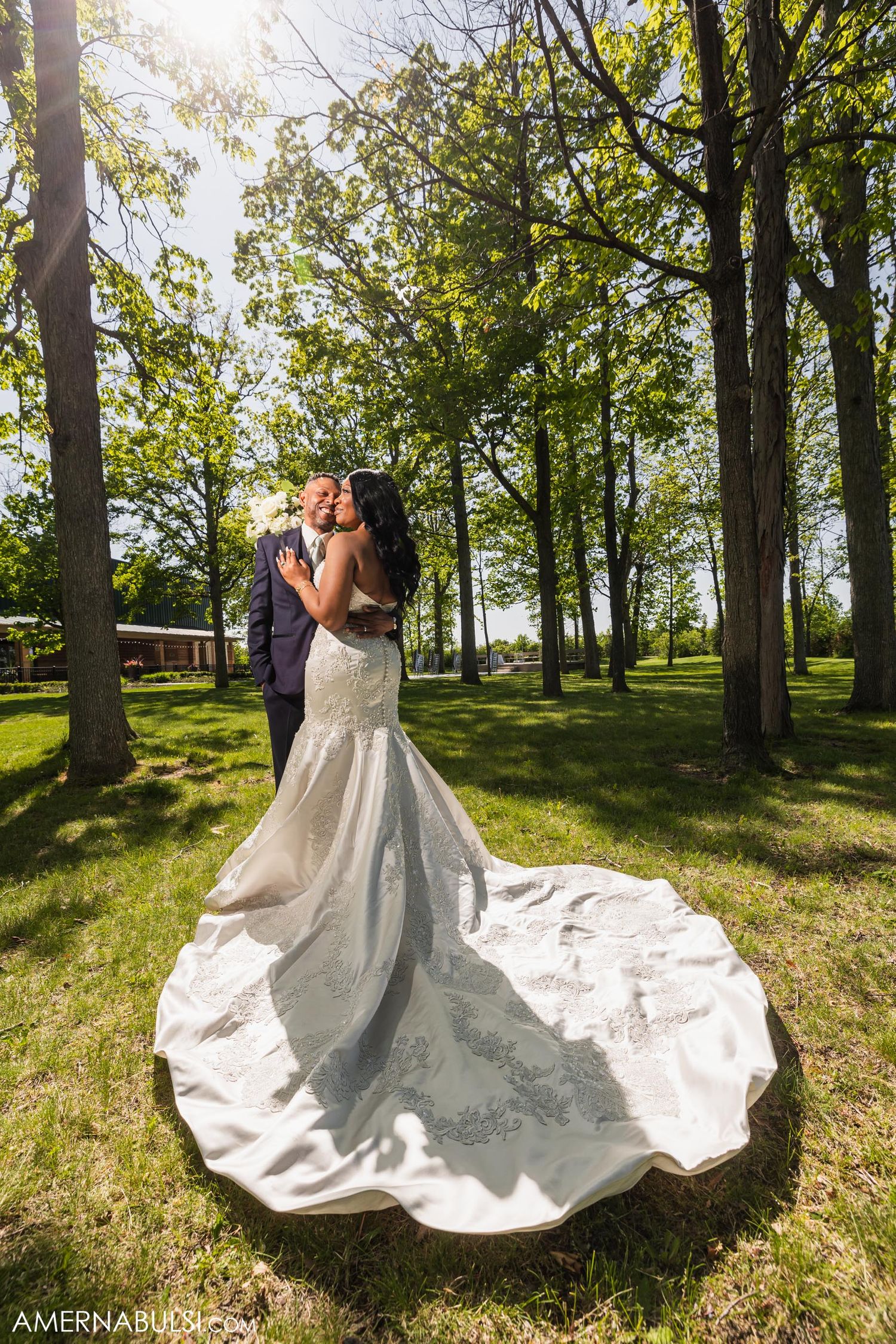 Wedding couple embraces in a sunlit forest with bride's dramatic train flowing behind.