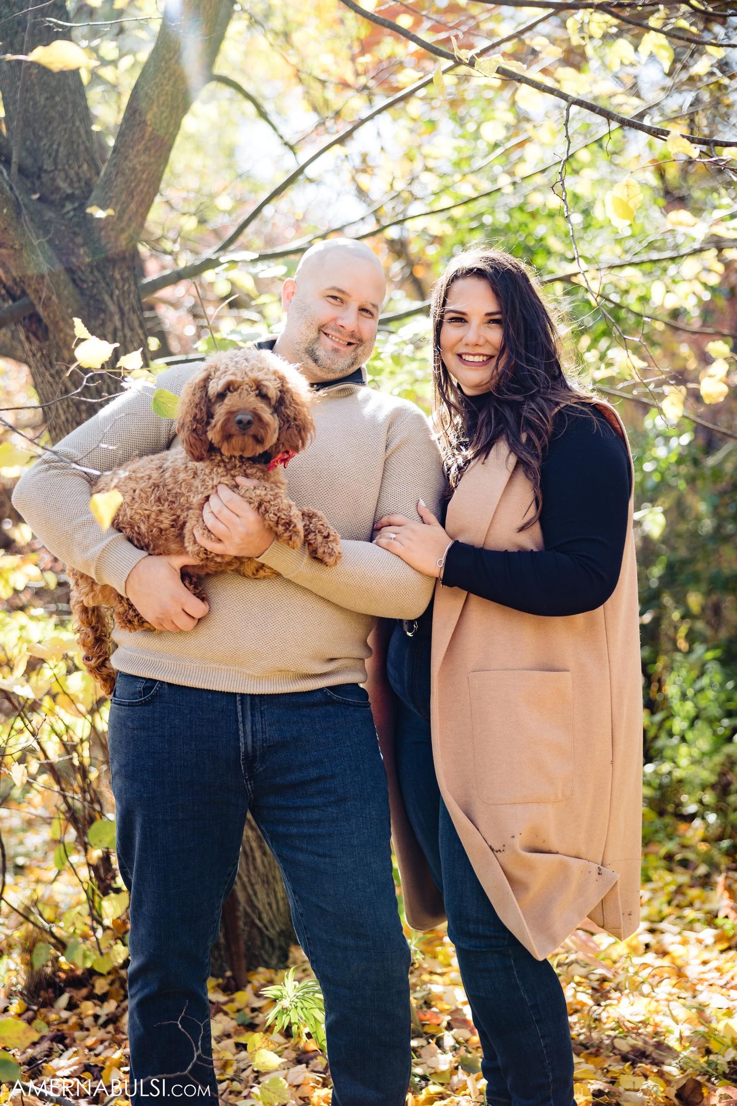Couple poses with their goldendoodle puppy among fall trees.