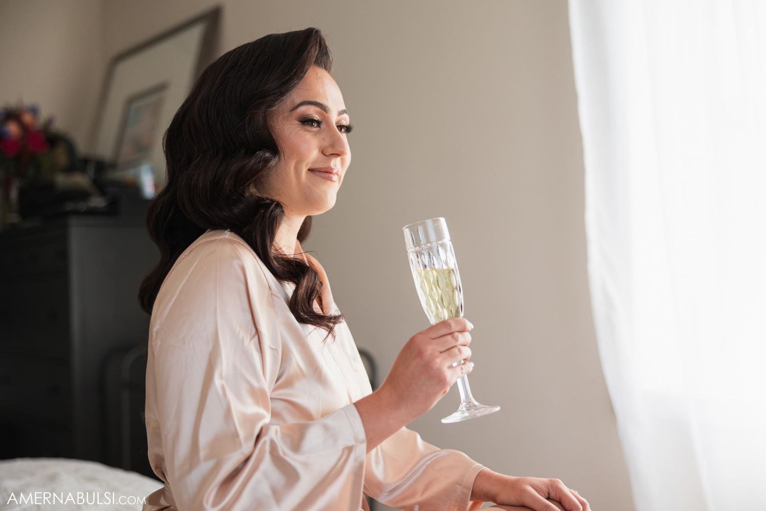 Woman in silk robe holding champagne glass by window during wedding preparations.