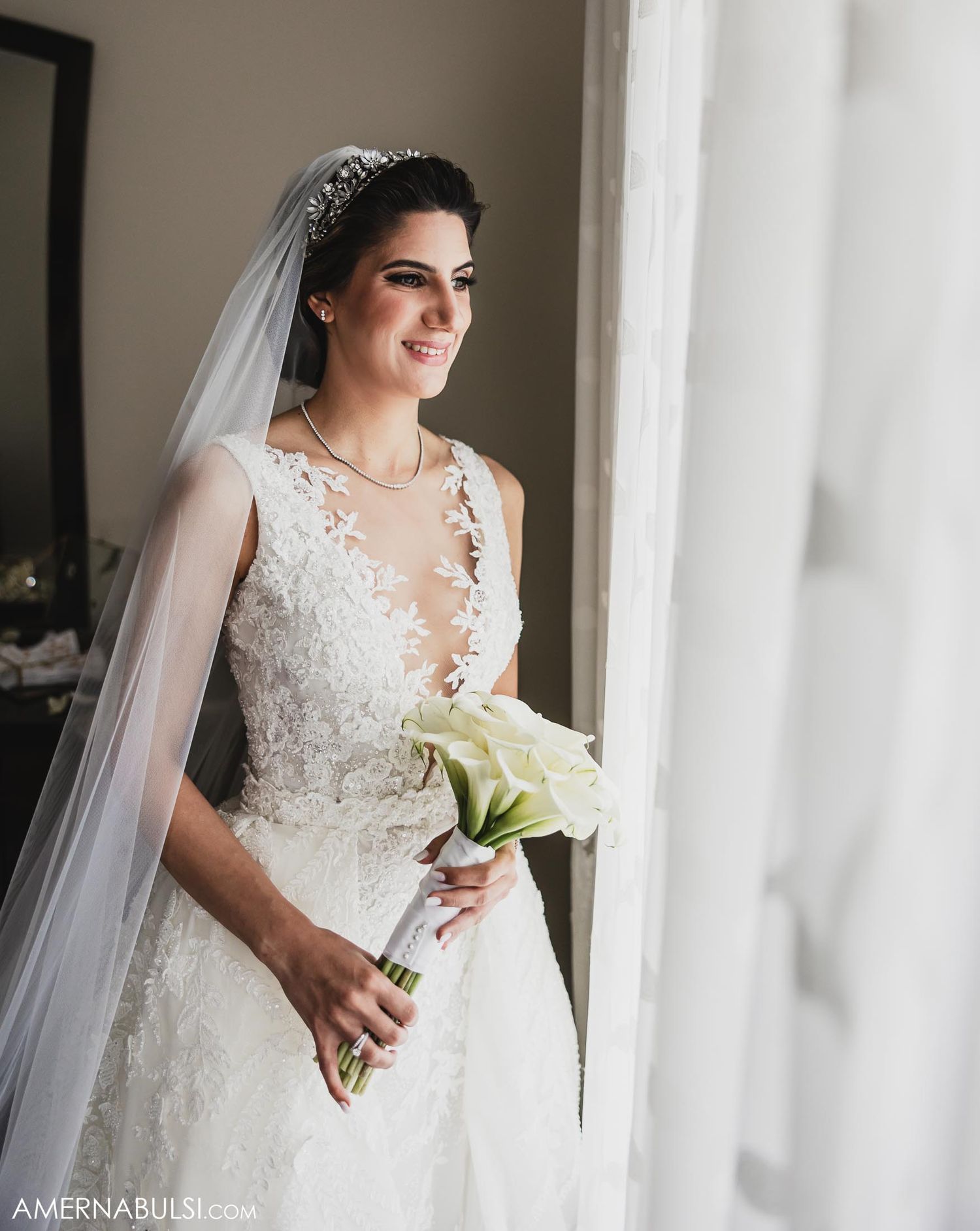 Bride in elegant lace gown holds white bouquet while standing by window.