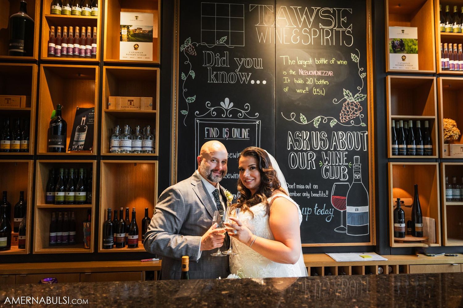 A couple toasts at a rustic bar with wooden shelves and chalkboard menu wall.