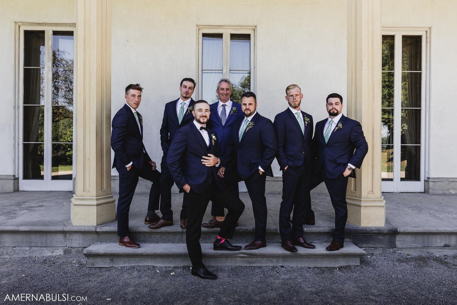 Groomsmen in navy suits pose on classic stone steps of elegant venue.