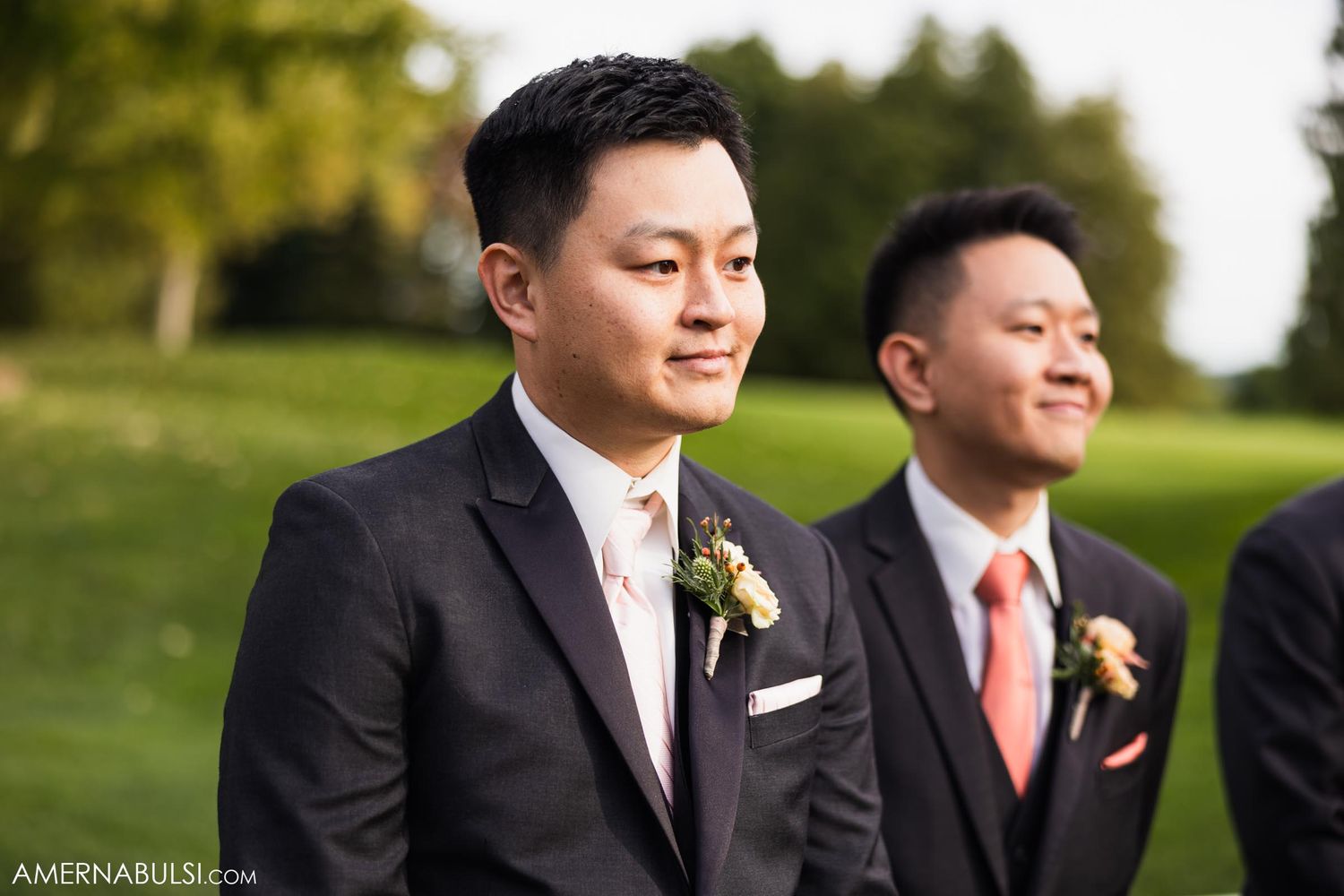 Groomsmen in black tuxedos stand together during an outdoor wedding ceremony.
