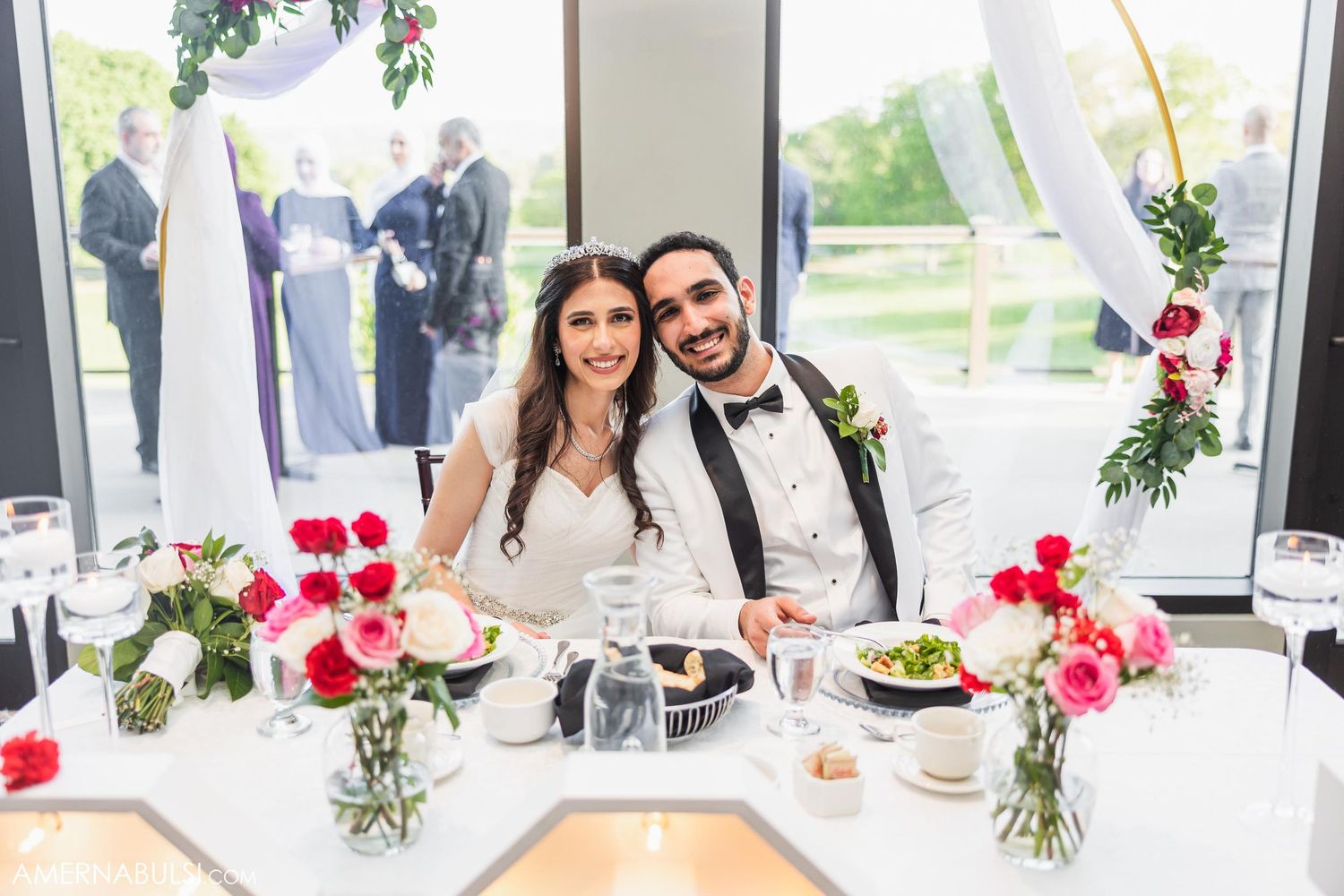 A couple sits at their sweetheart table decorated with red roses at their wedding reception.