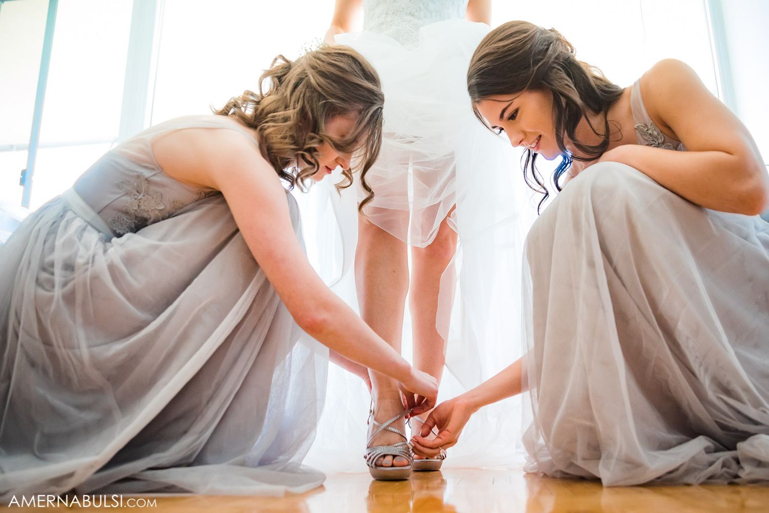 Two bridesmaids in gray dresses kneel together helping with wedding preparations.