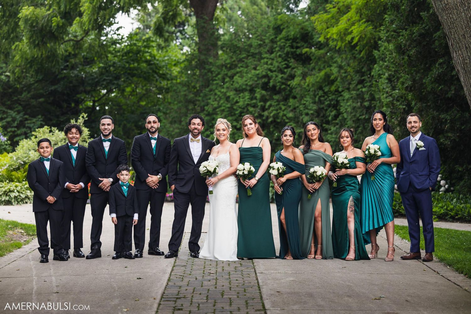 Wedding party poses on stone pathway with bridesmaids in green dresses and groomsmen in black suits.