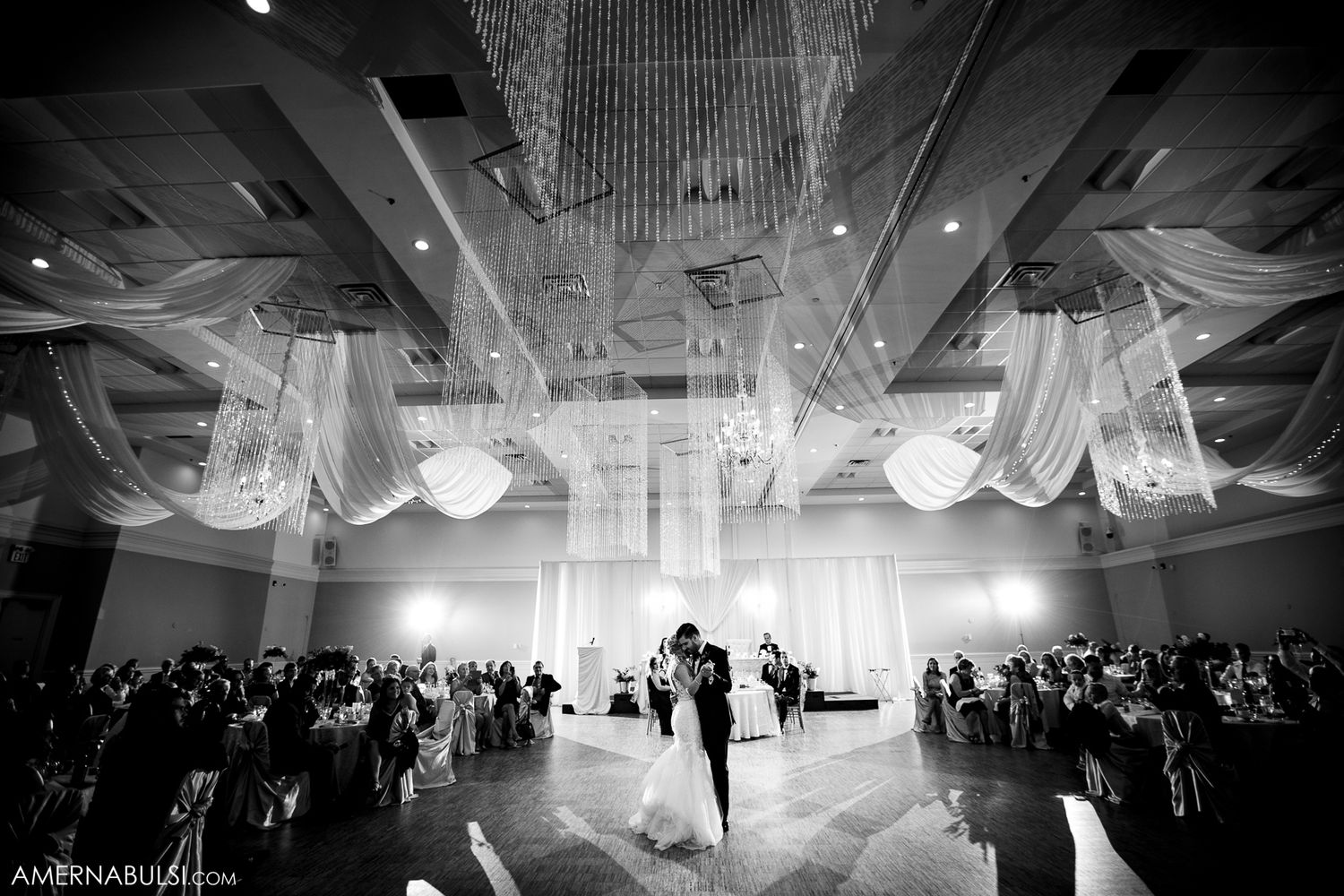 Wedding reception dance floor filled with guests under elegant white drapery and chandeliers during evening celebration.
