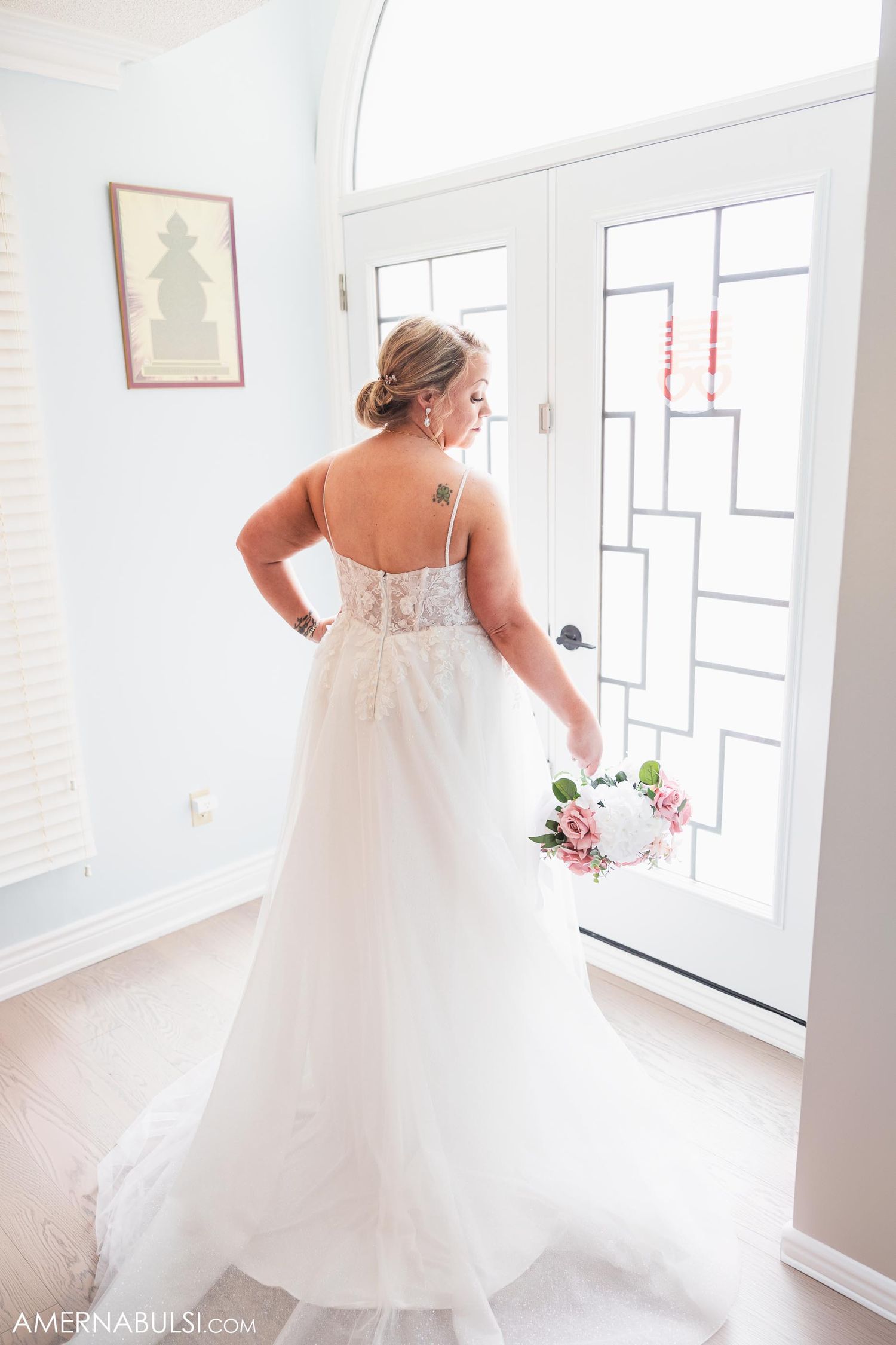 Back view of a bride in a flowing white wedding gown standing by a modern glass door.