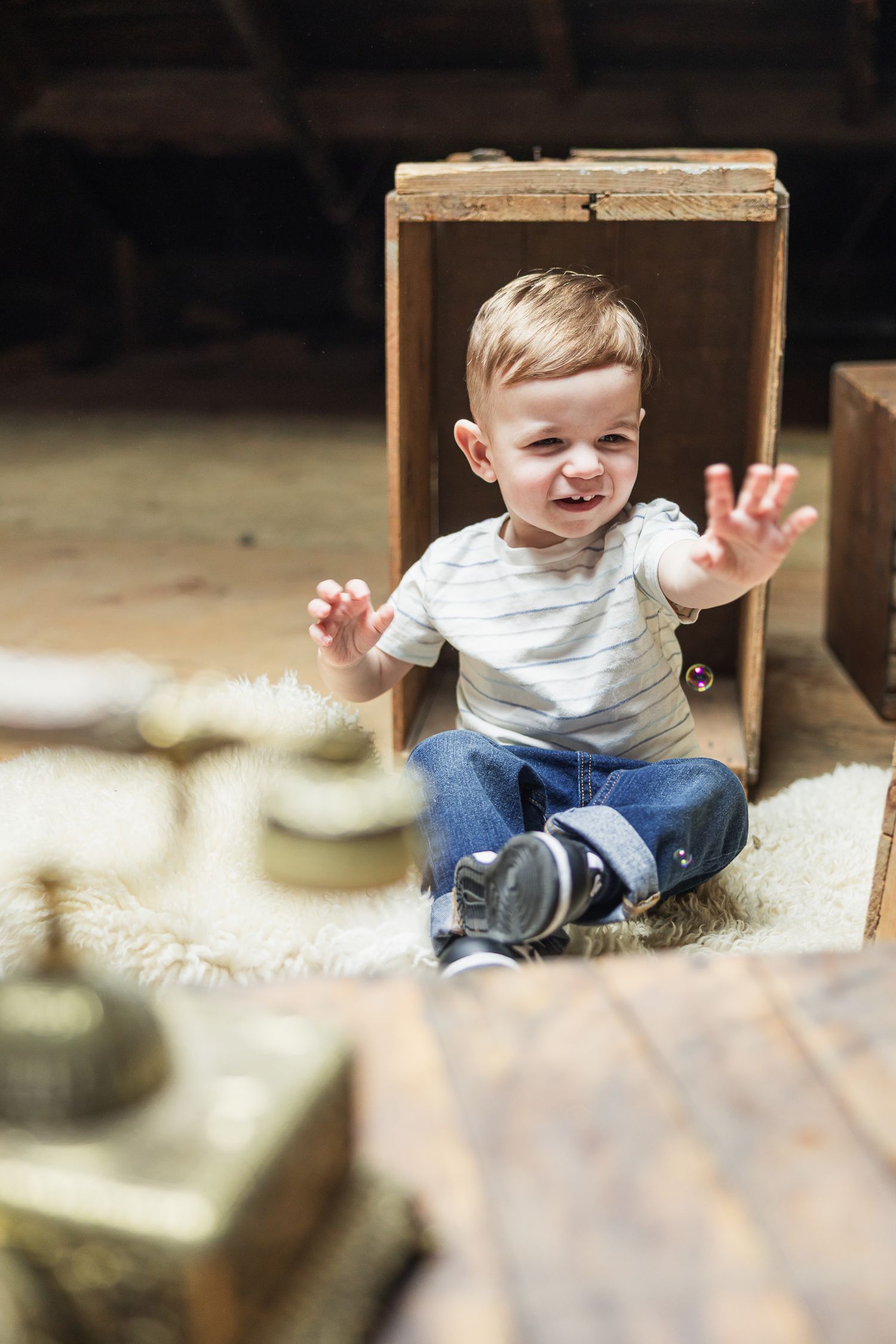 A toddler wearing a white shirt and jeans sits playfully in a wooden box with outstretched hands.