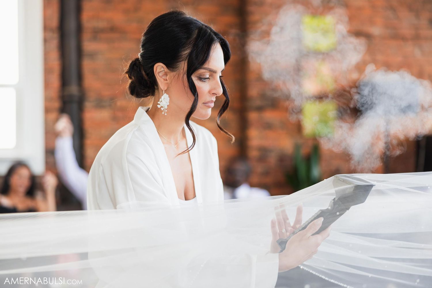 Woman in white robe examines flowing veil with steam in rustic brick setting.