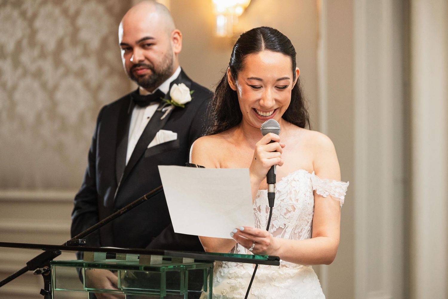 A bride reads from papers at a podium during a wedding speech while her groom stands beside her.