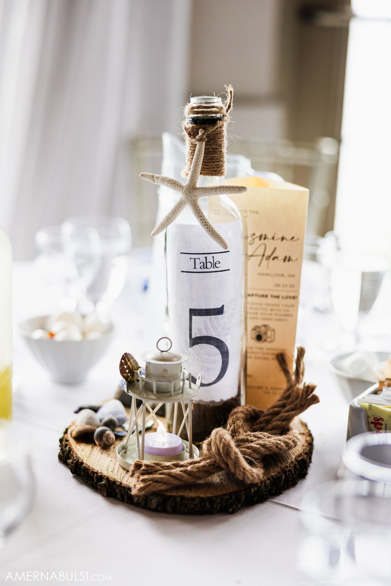 Beach themed wedding table centerpiece featuring a bottle with number 5, starfish, and nautical rope on a wooden base.