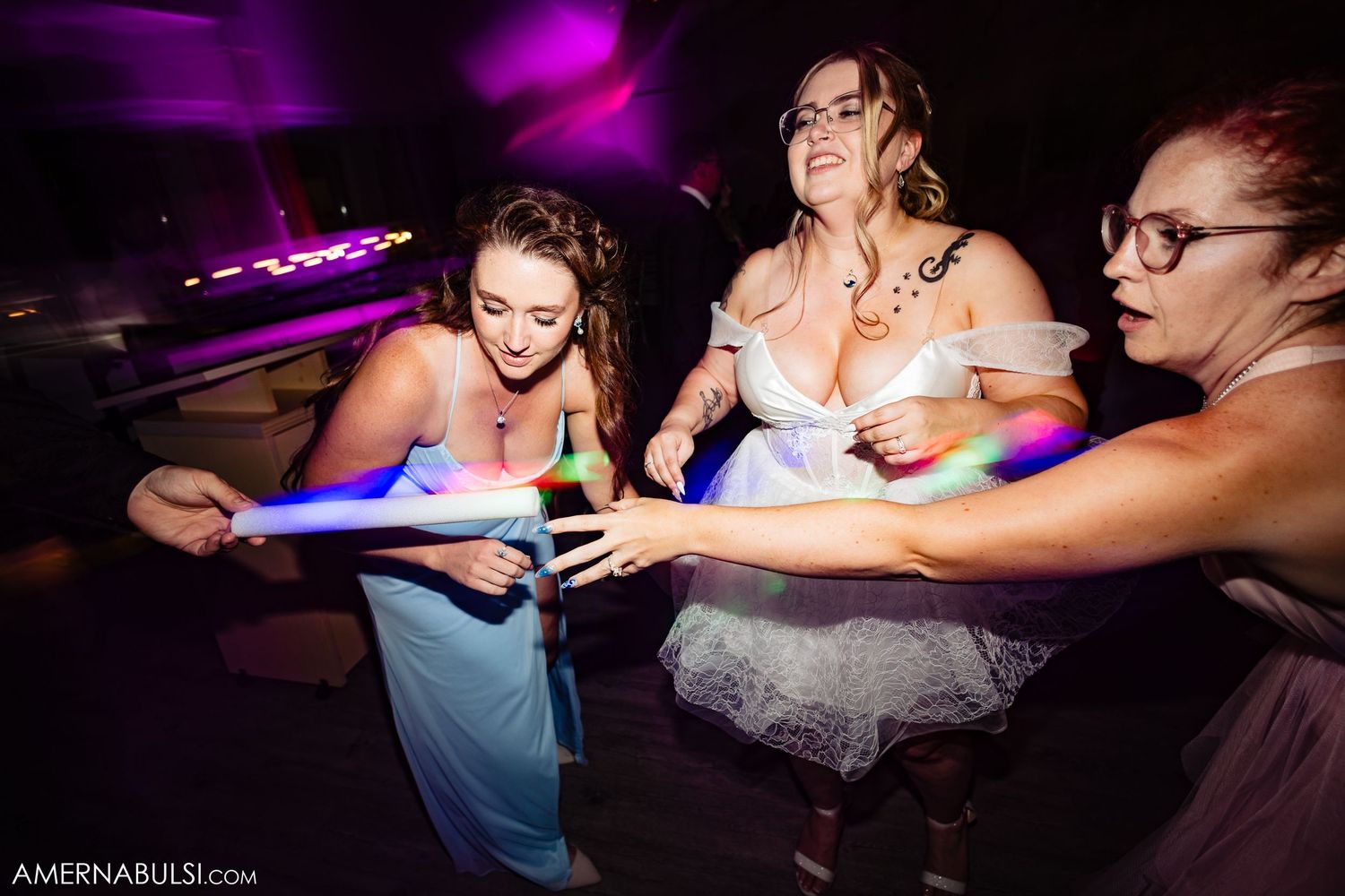 Guests lean across a table at a dimly lit party venue with purple lighting and dance floor atmosphere.