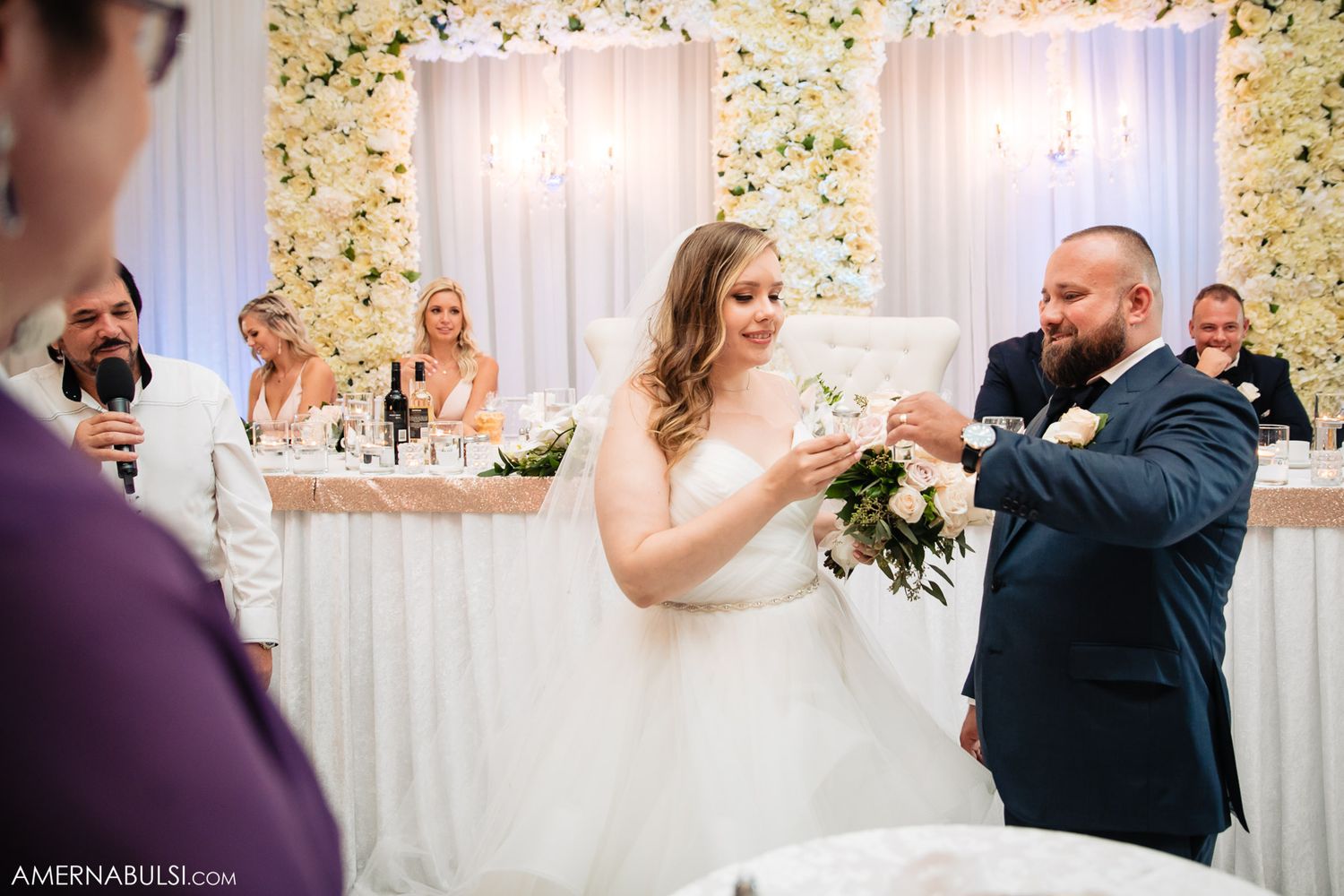 Wedding celebration in a white and gold decorated venue with floral arrangements and elegant lighting.