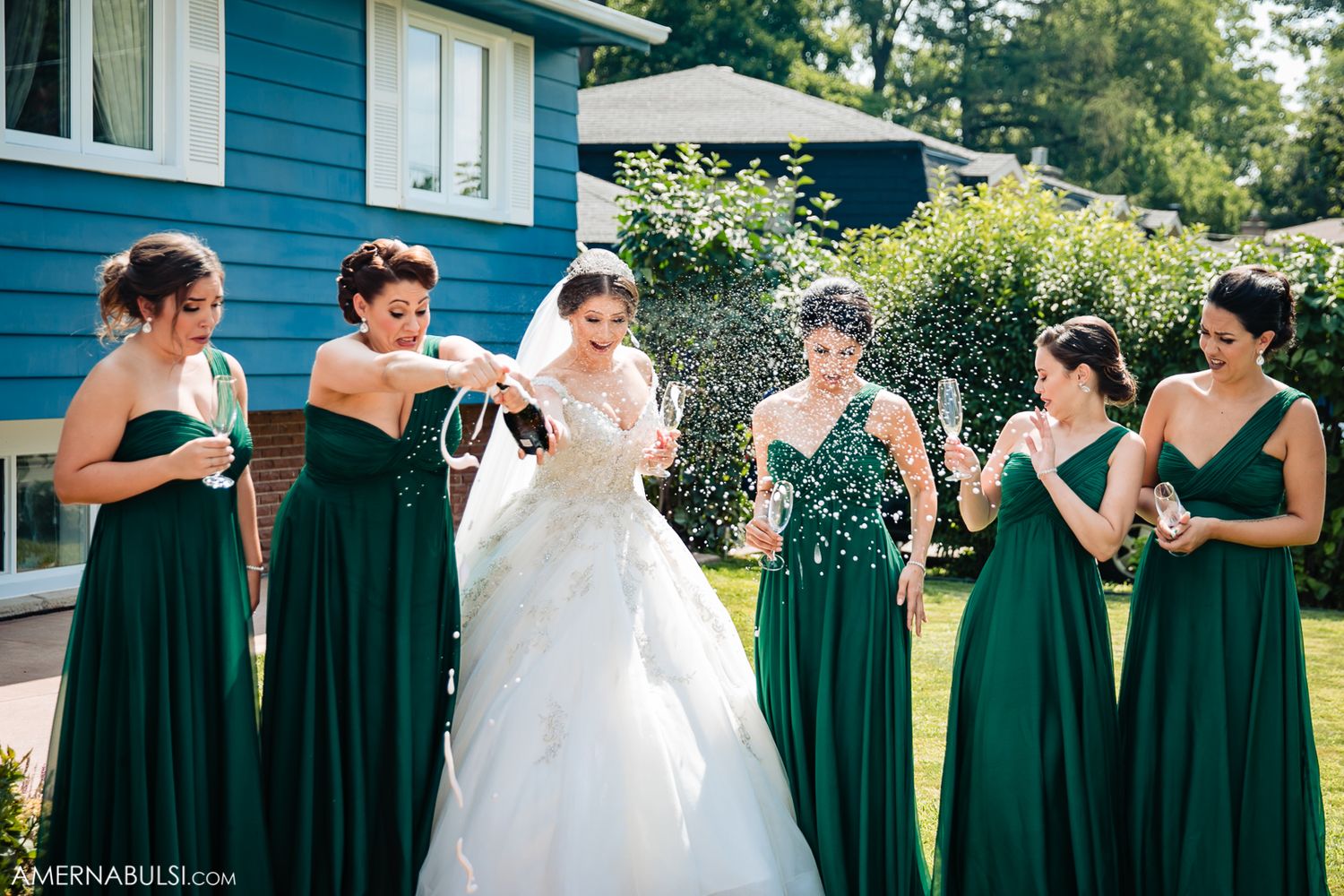 Wedding party in emerald green dresses and white gown posed in front of blue house.
