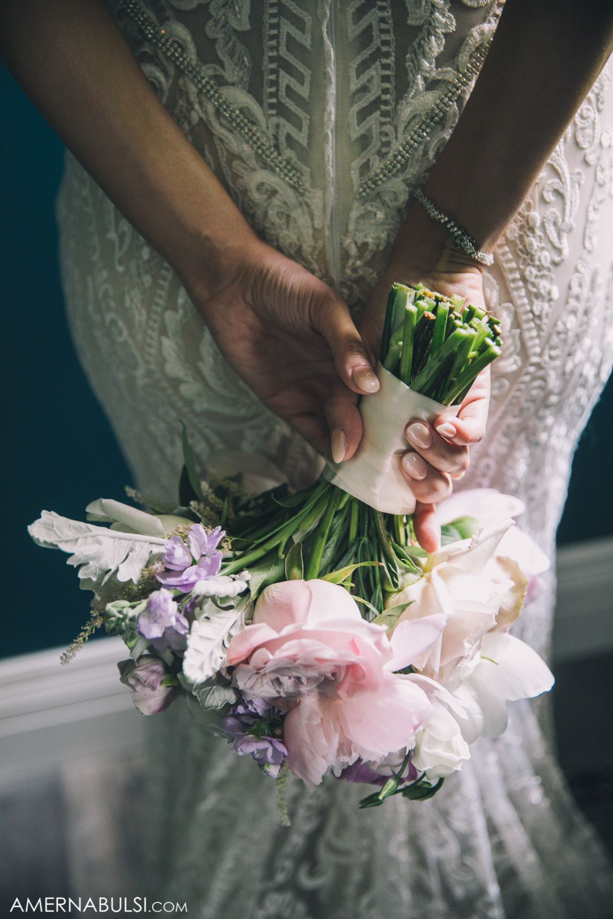 Close-up of hands holding a delicate bridal bouquet with pink and purple flowers against a white lace dress.