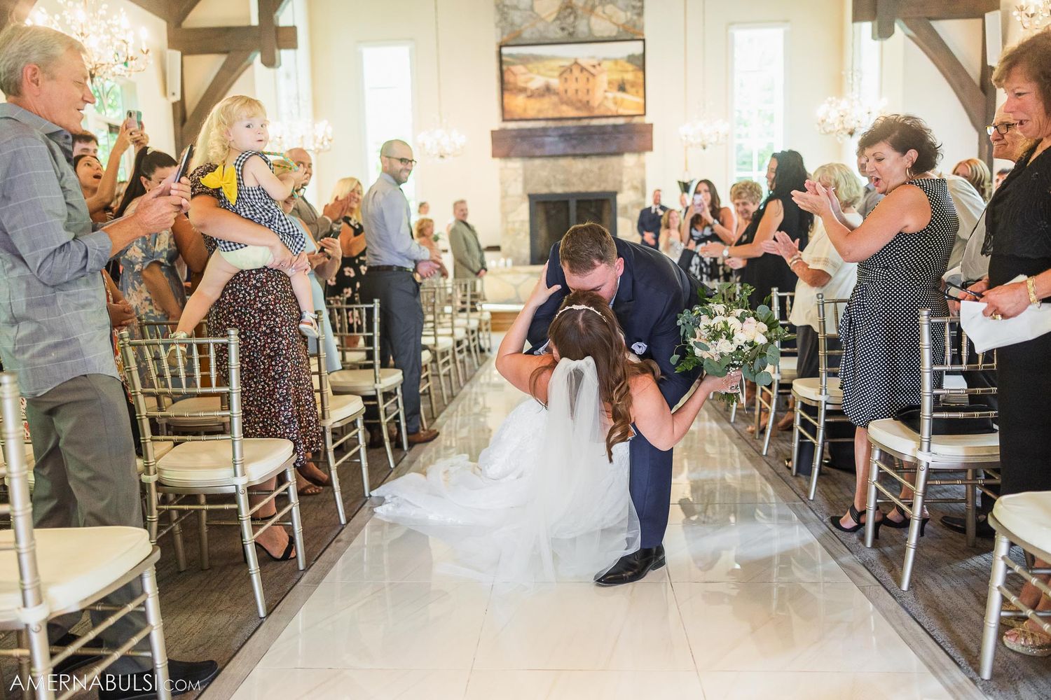 Wedding couple shares romantic dip kiss in church aisle while guests capture the moment on smartphones.