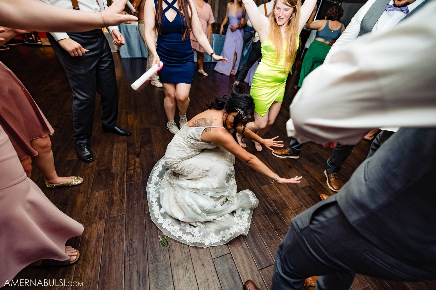 Wedding guests dancing energetically on a wooden dance floor with a person in a white dress crouched down.