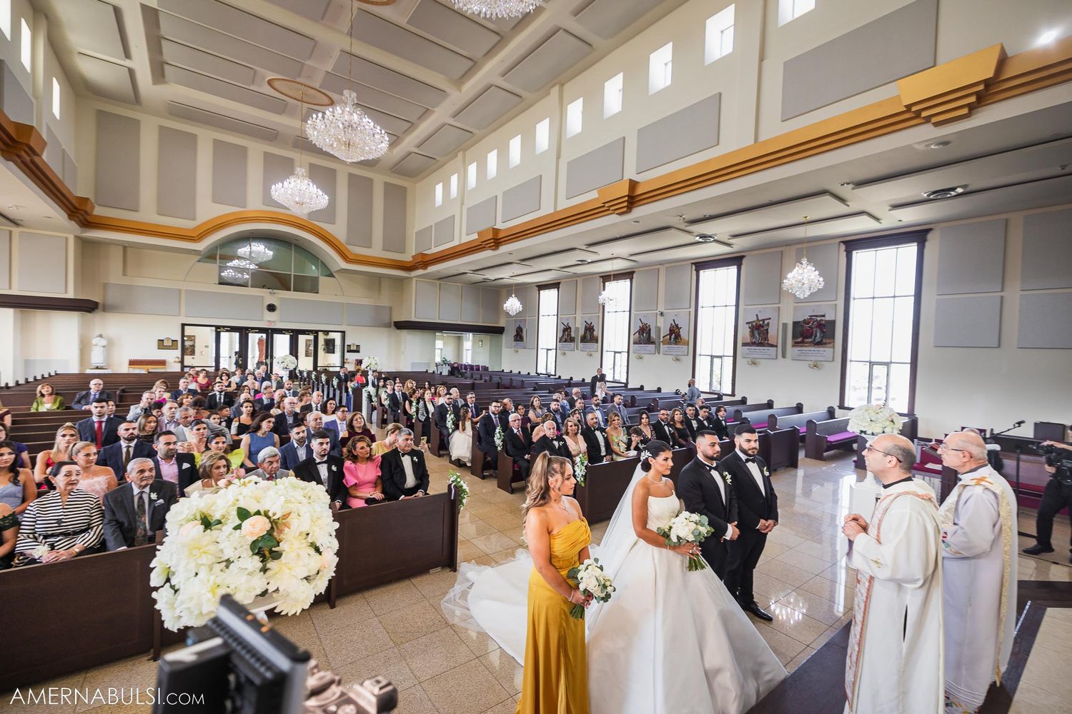 Elegant Catholic church wedding ceremony with large crowd gathered in ornate hall with high ceilings and chandelier.