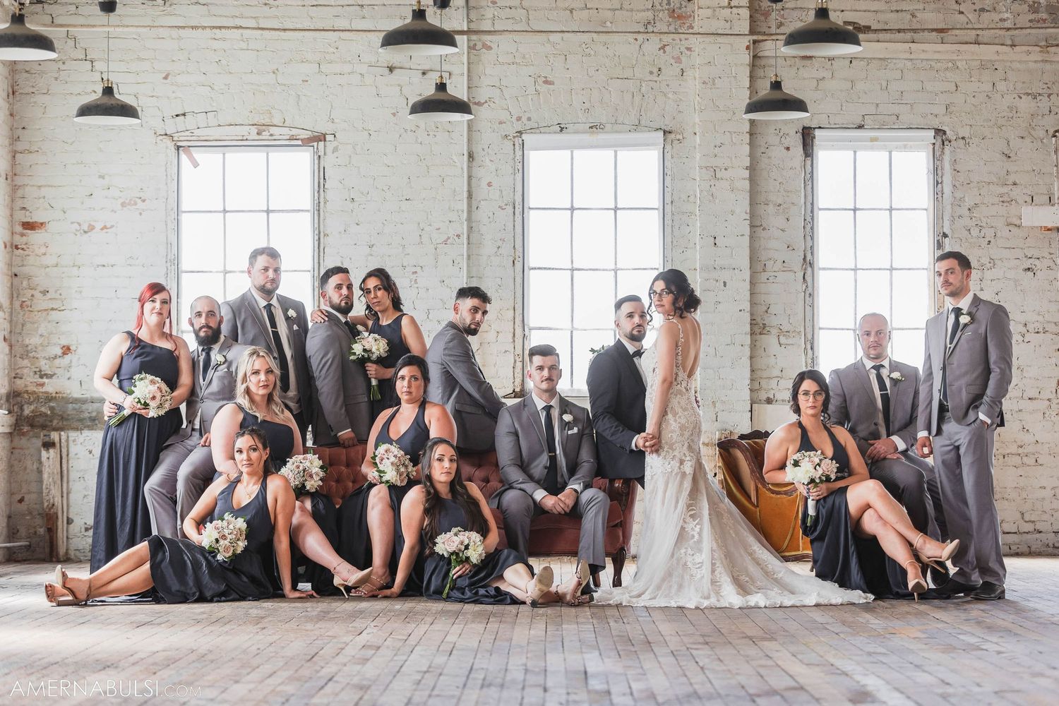 Wedding party poses together in industrial loft space with white brick walls and large windows.