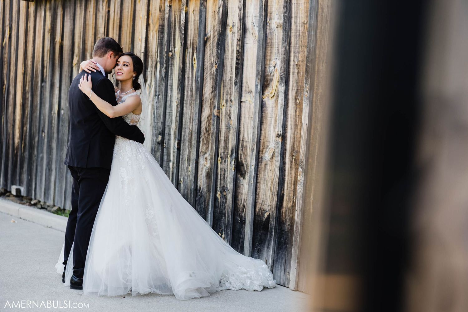 Wedding couple embraces against rustic wooden wall with bride wearing flowing white gown.