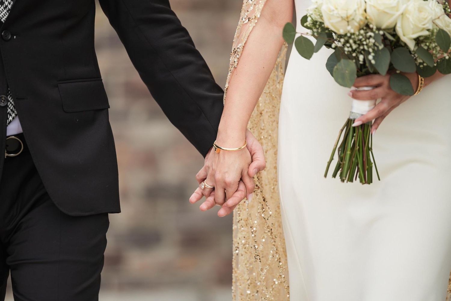 Close-up of couple holding hands with white bouquet and gold dress details.