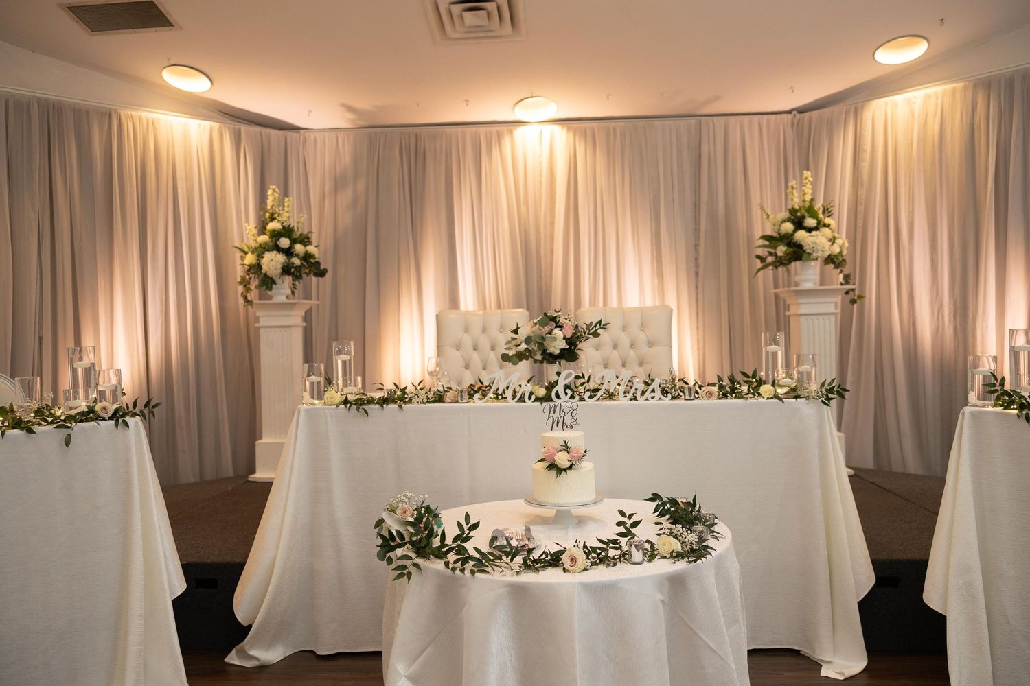 Elegant wedding reception setup with white draped backdrop, floral arrangements, and illuminated head table decor.