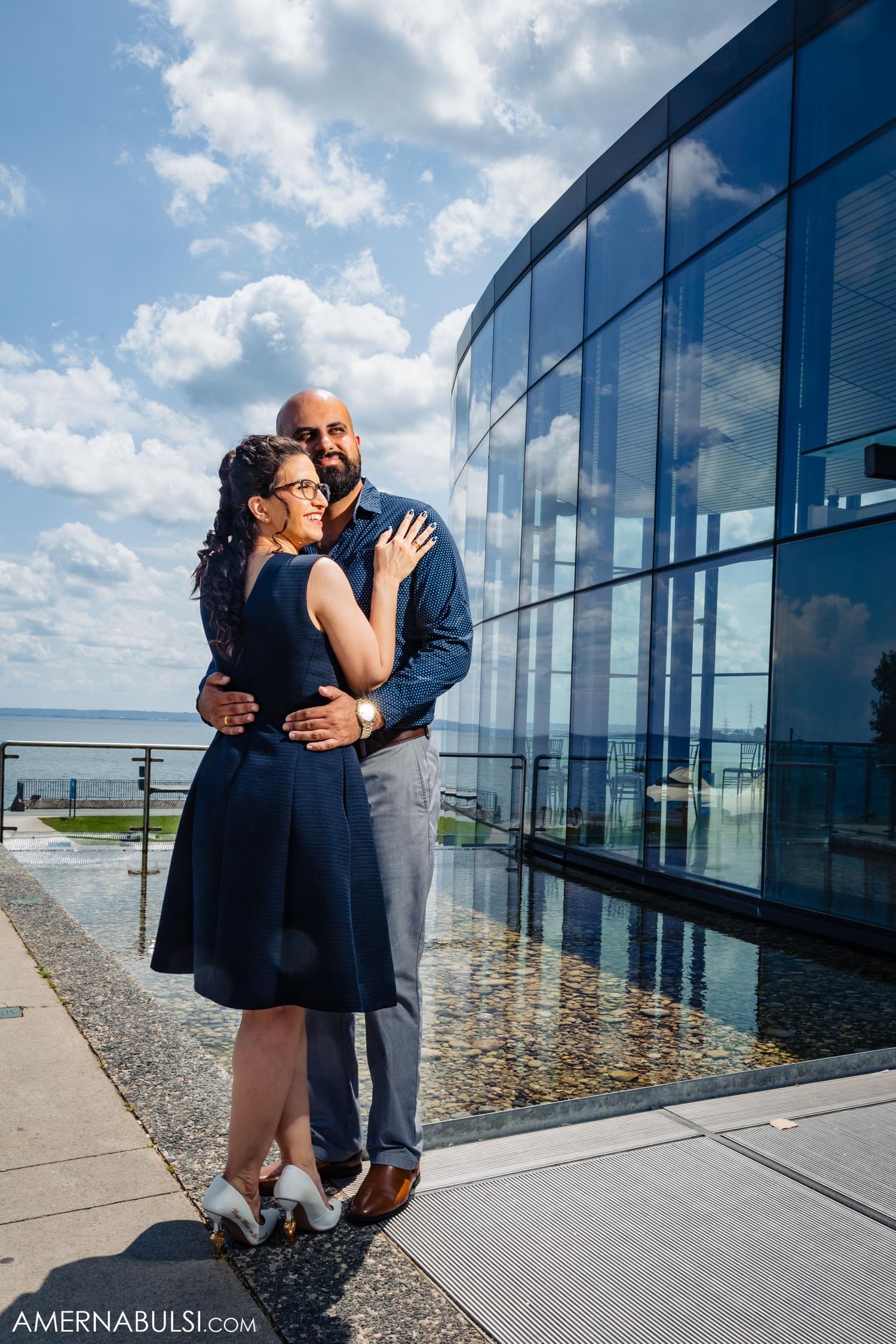 A couple poses in front of a modern glass building with water views.