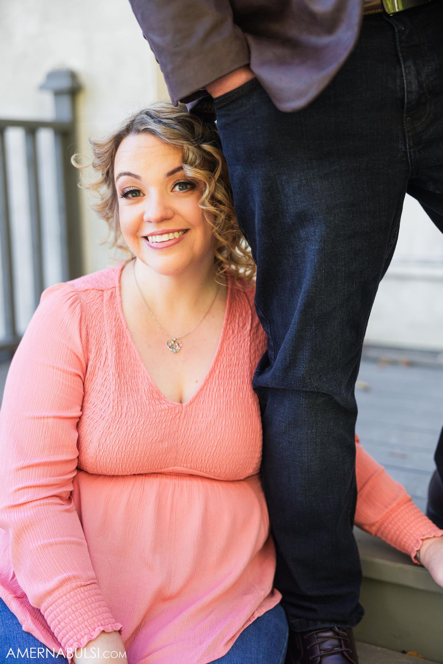 A woman in a coral pink sweater smiles warmly while sitting on a porch.