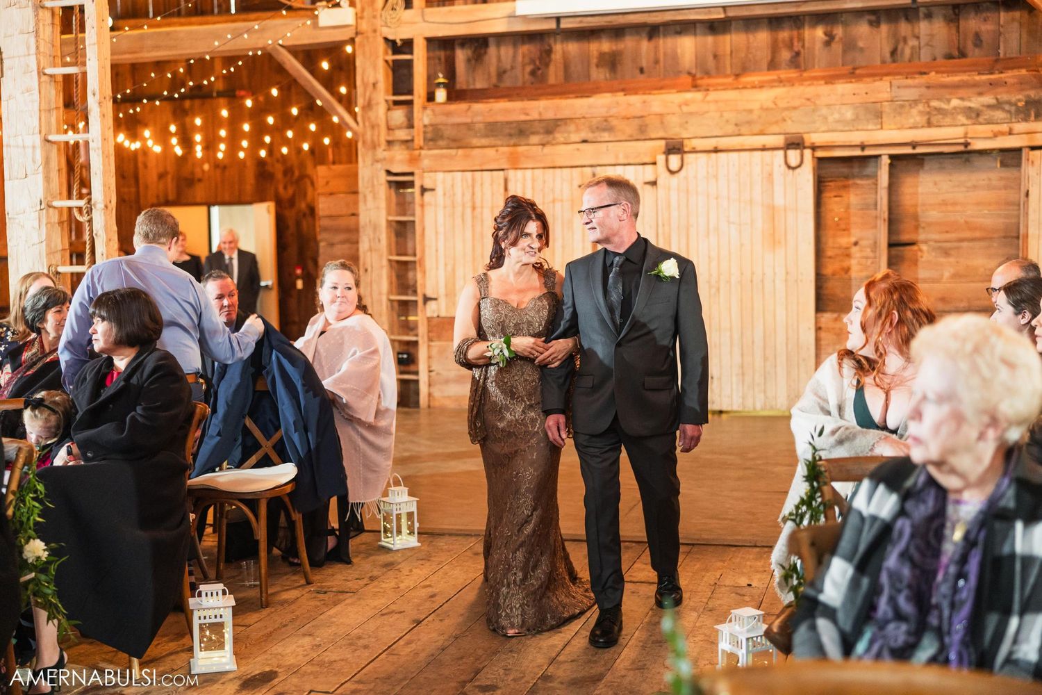 Wedding couple walks down aisle in rustic barn venue lit with string lights and lantern decorations.