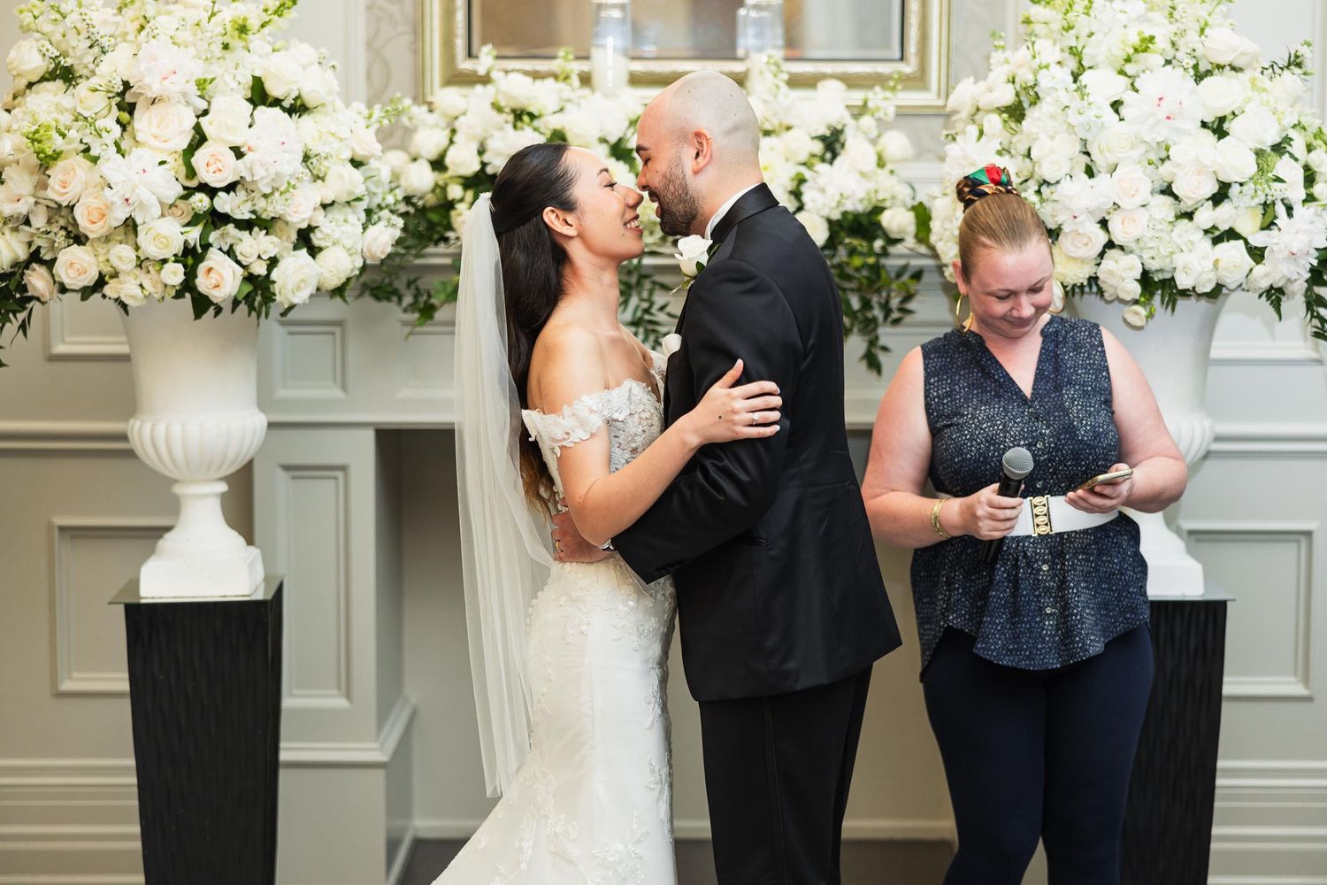 Wedding ceremony in elegant room with white floral arrangements and gilded mirrors.