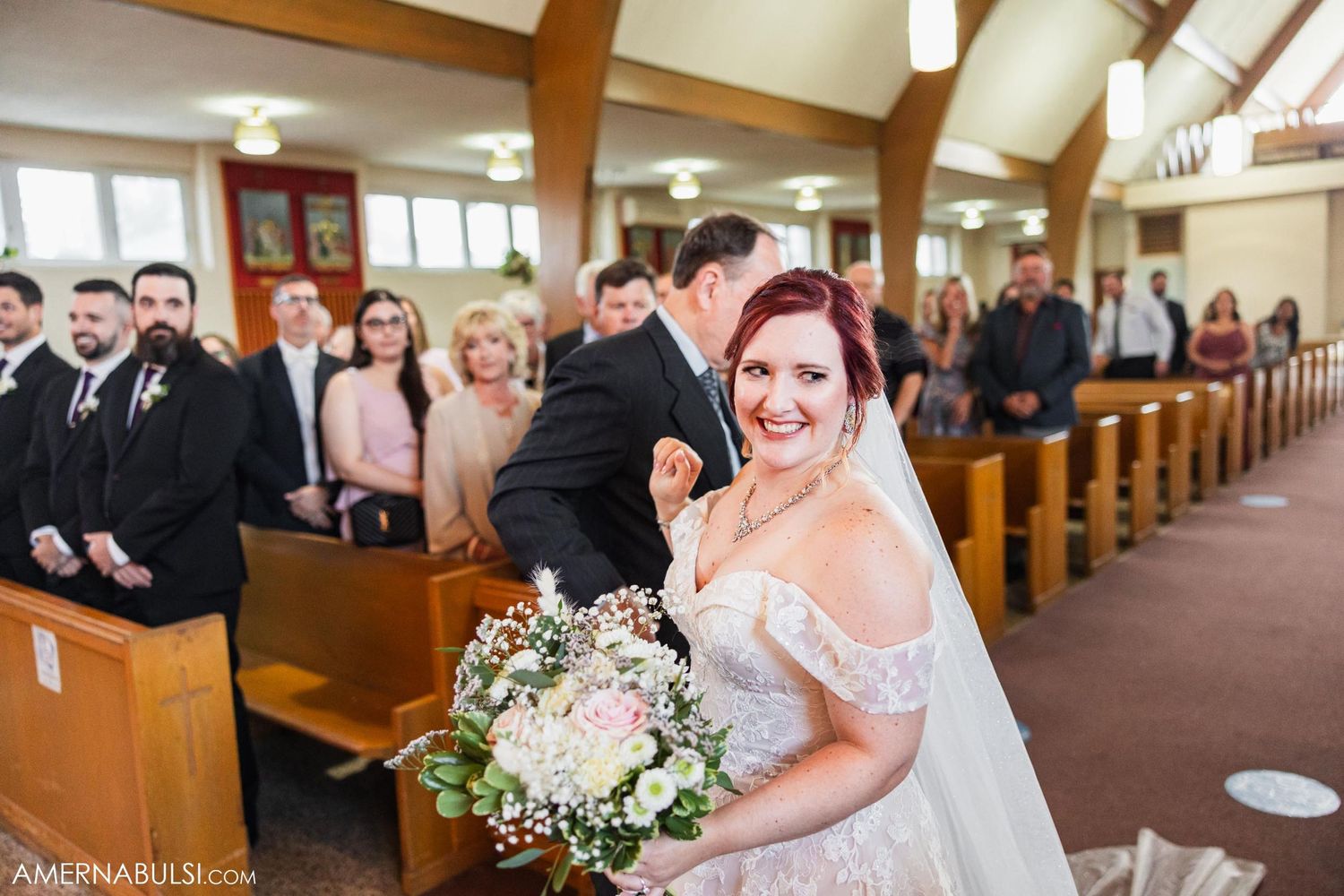 Bride holding floral bouquet while walking down church aisle with wooden pews.