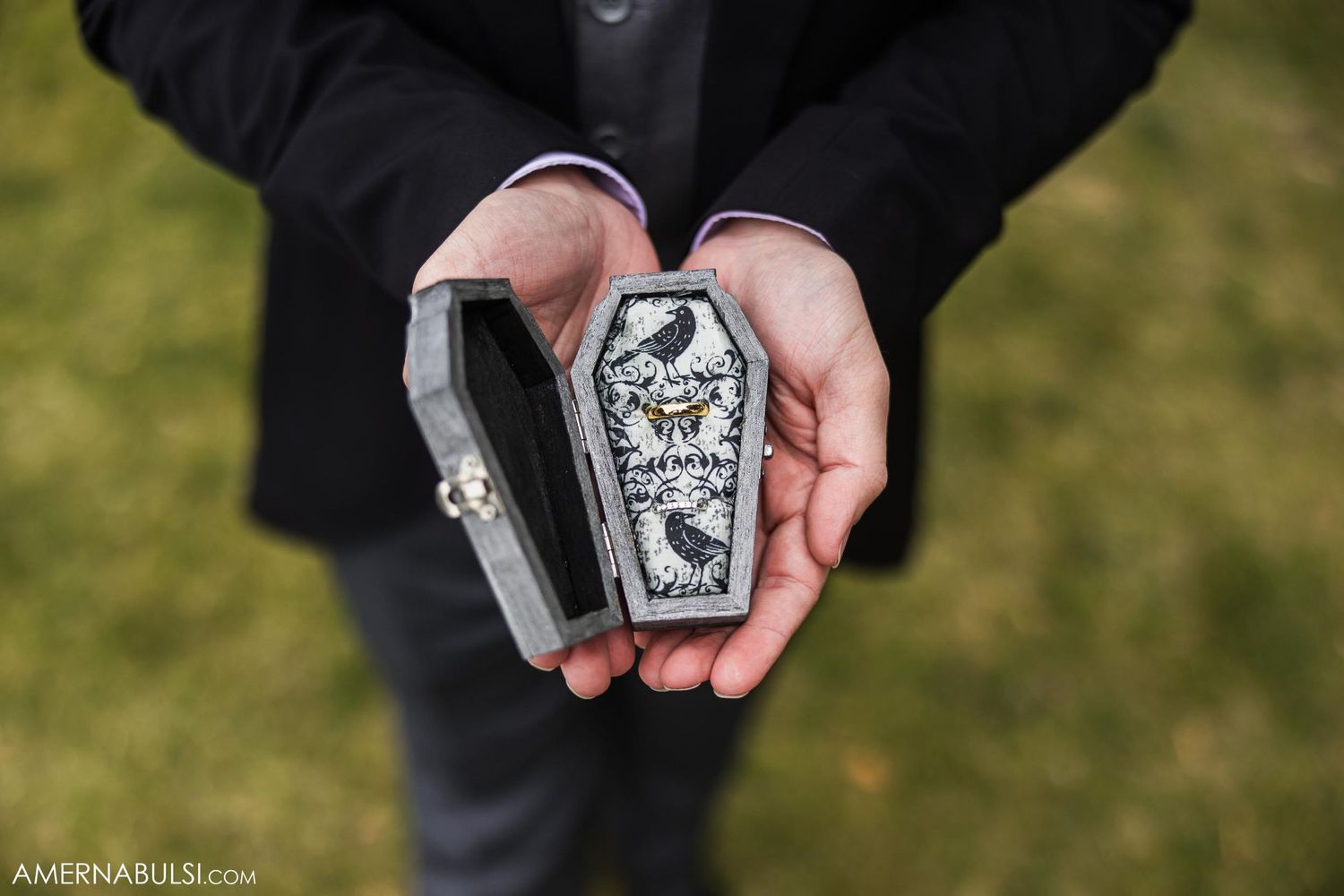 Hands holding a decorative silver ring box against dark fabric outdoors.