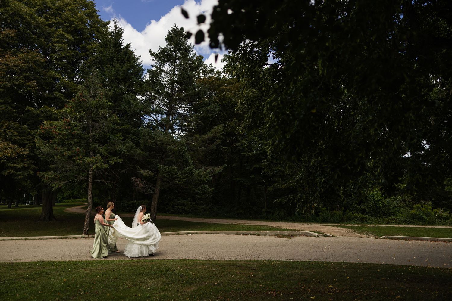Bride in white gown with attendant carrying train in lush green park setting on wedding day.