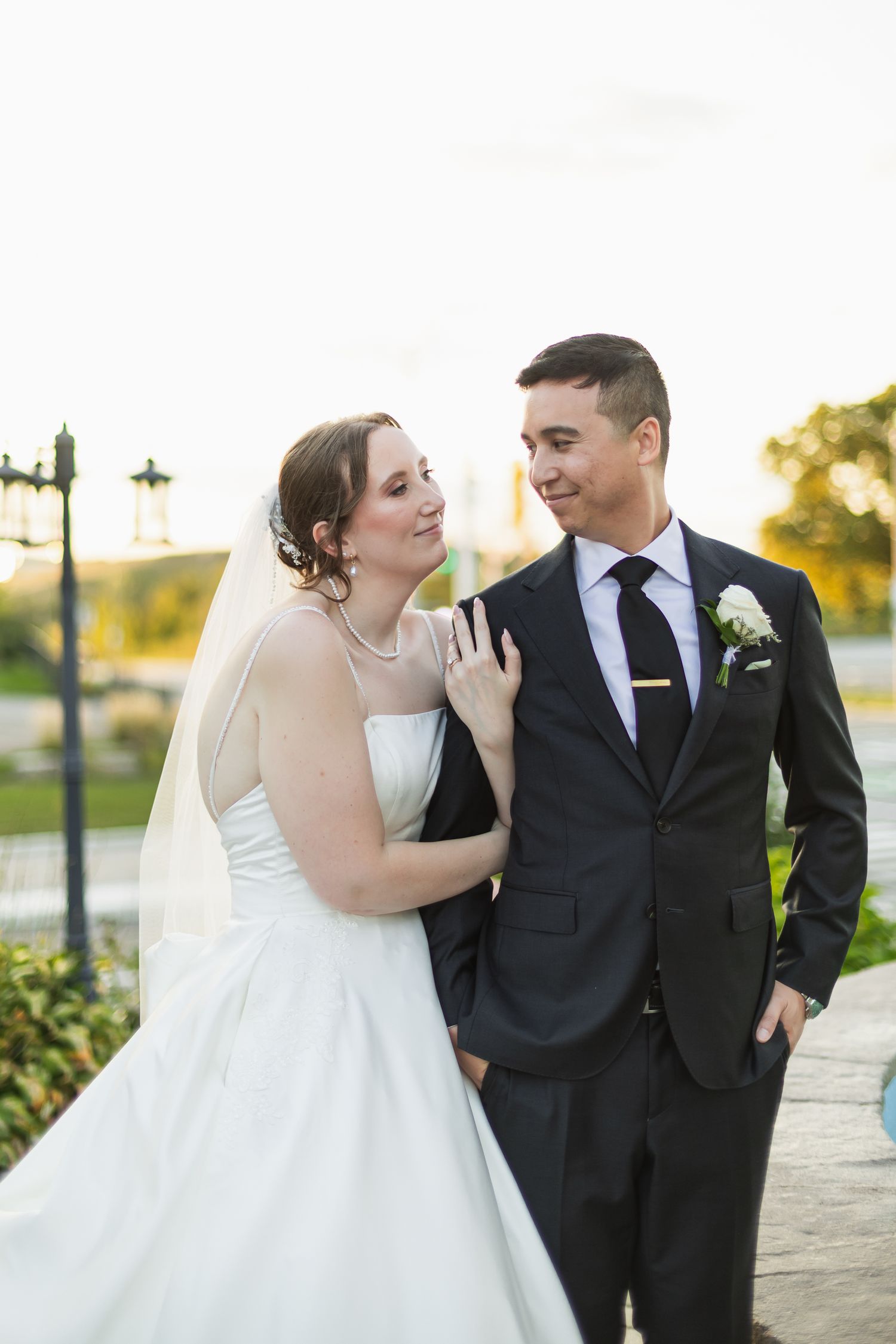 Bride in white gown and veil smiles at groom in dark suit with boutonniere during outdoor wedding portrait.