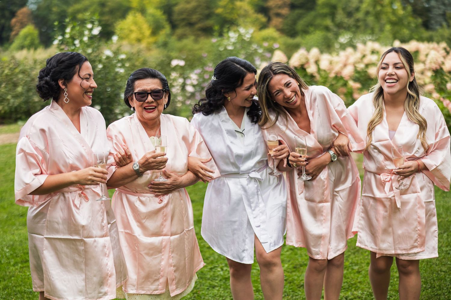 Bridesmaids in pink satin robes laughing and holding champagne glasses in a garden before wedding.