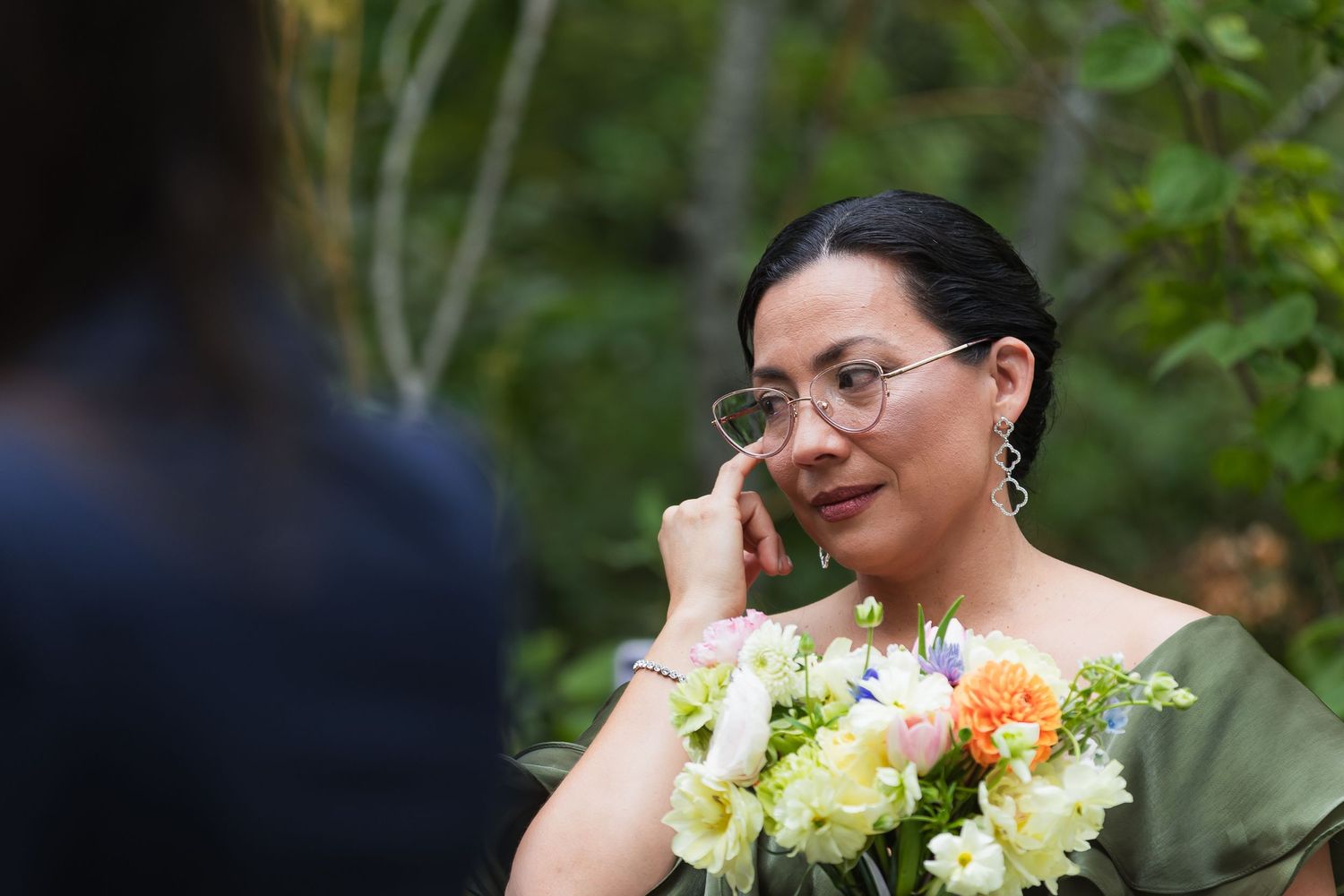 Emotional wedding guest wiping tears while holding a bouquet of white and peach flowers during an outdoor ceremony.