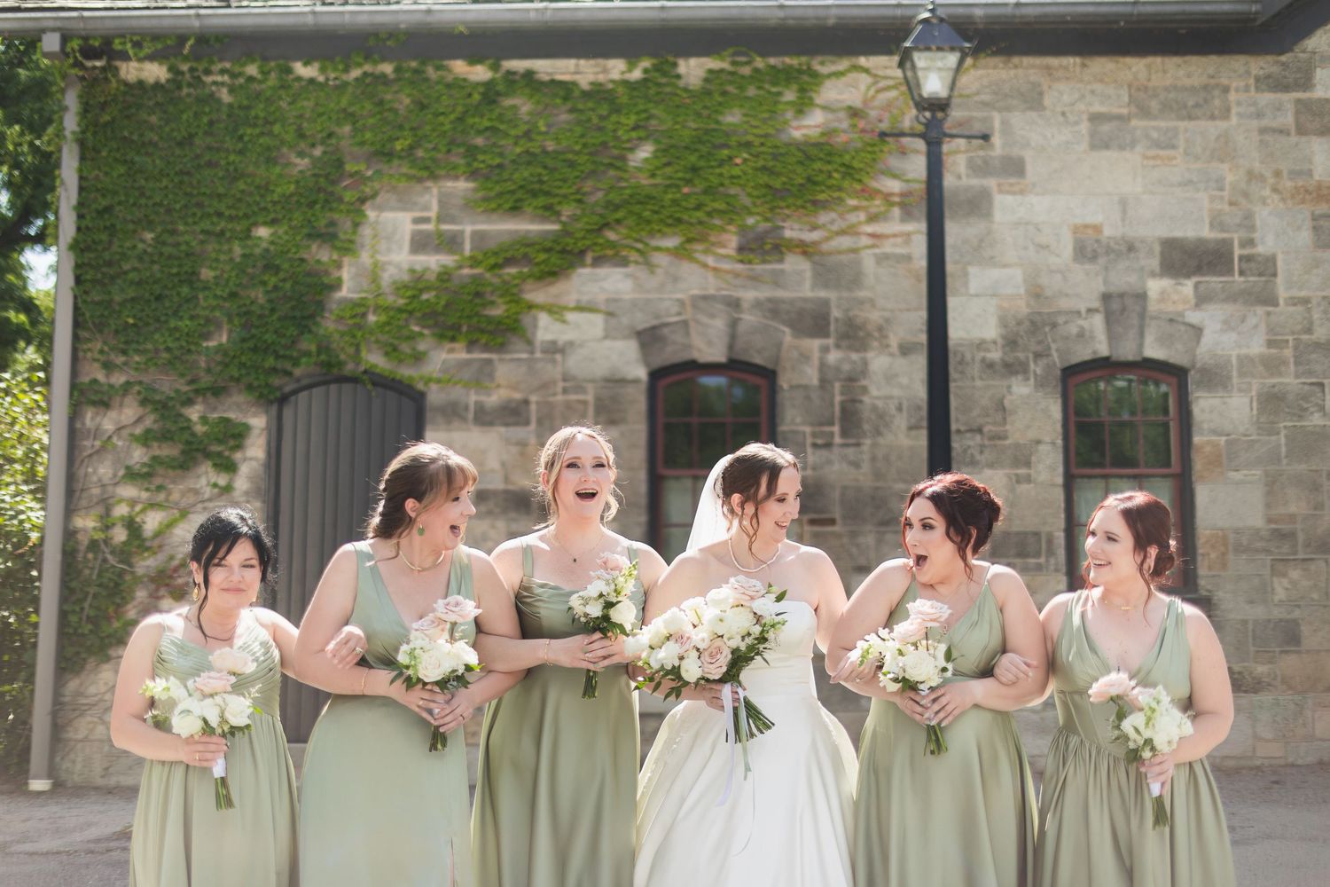 Bride in white gown stands with six bridesmaids in sage green dresses holding bouquets outside a stone building.