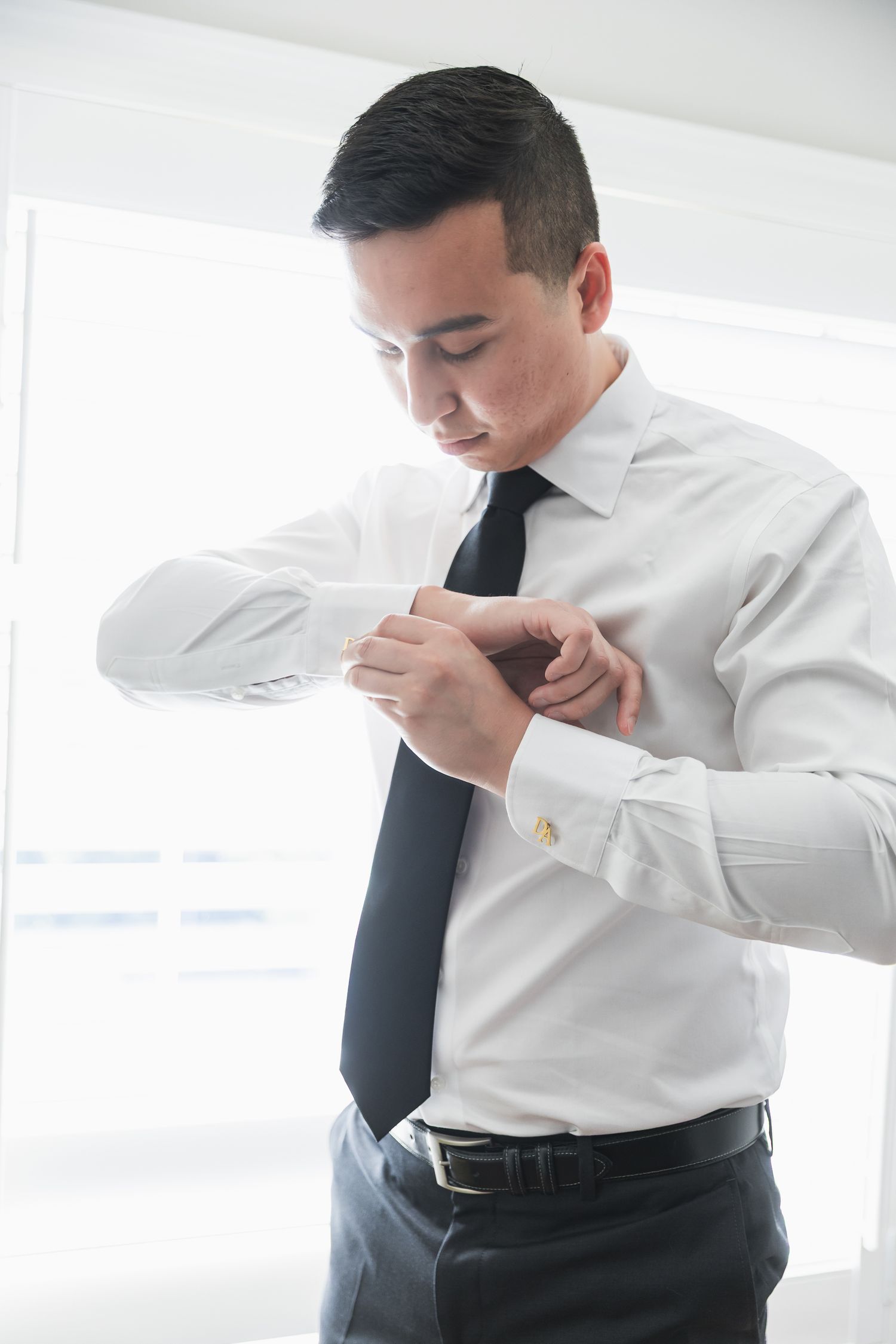 Groom in white shirt and black tie fastening cufflinks while getting dressed for wedding.