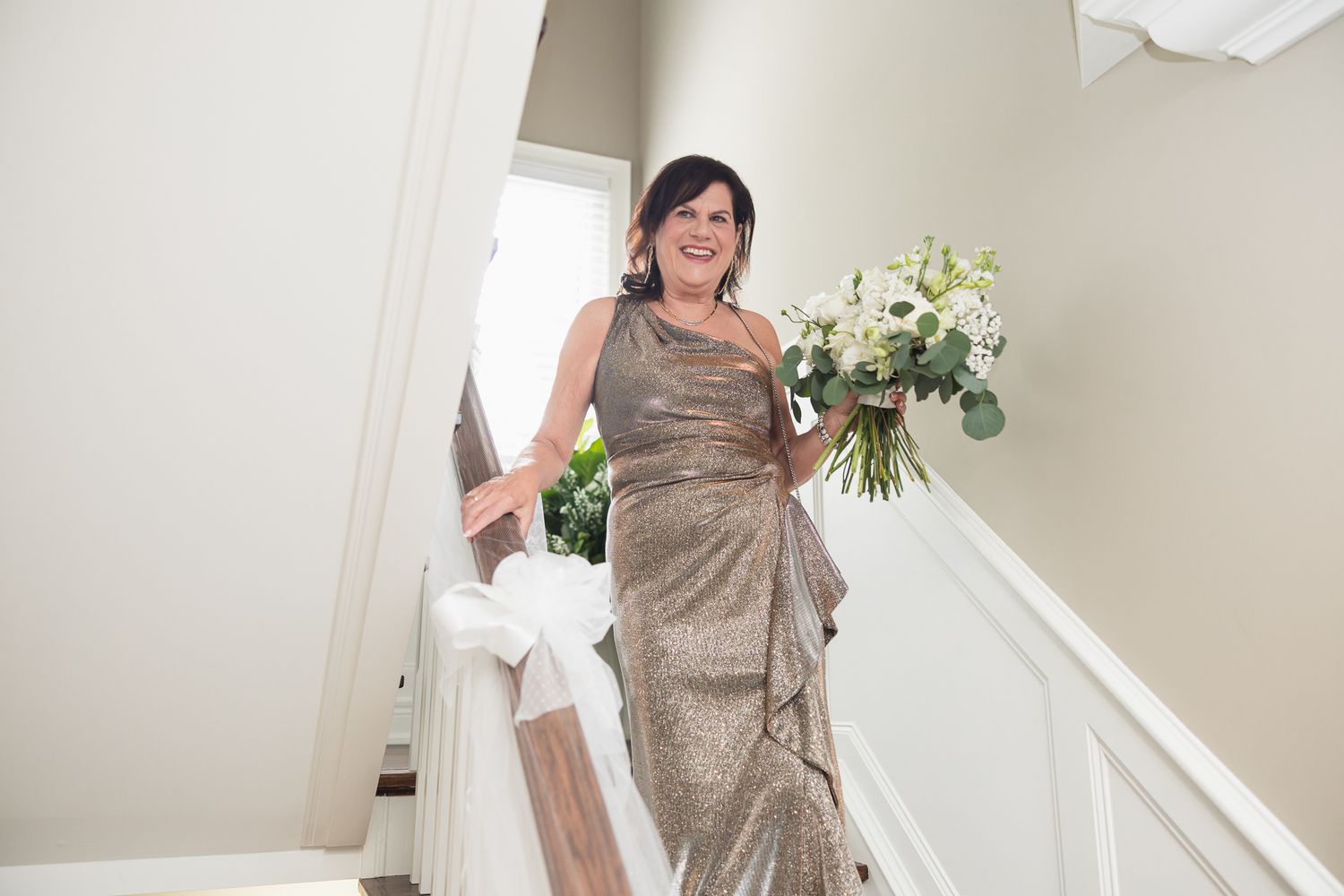 Woman in gold gown descending staircase holding white floral bouquet, smiling at wedding celebration.