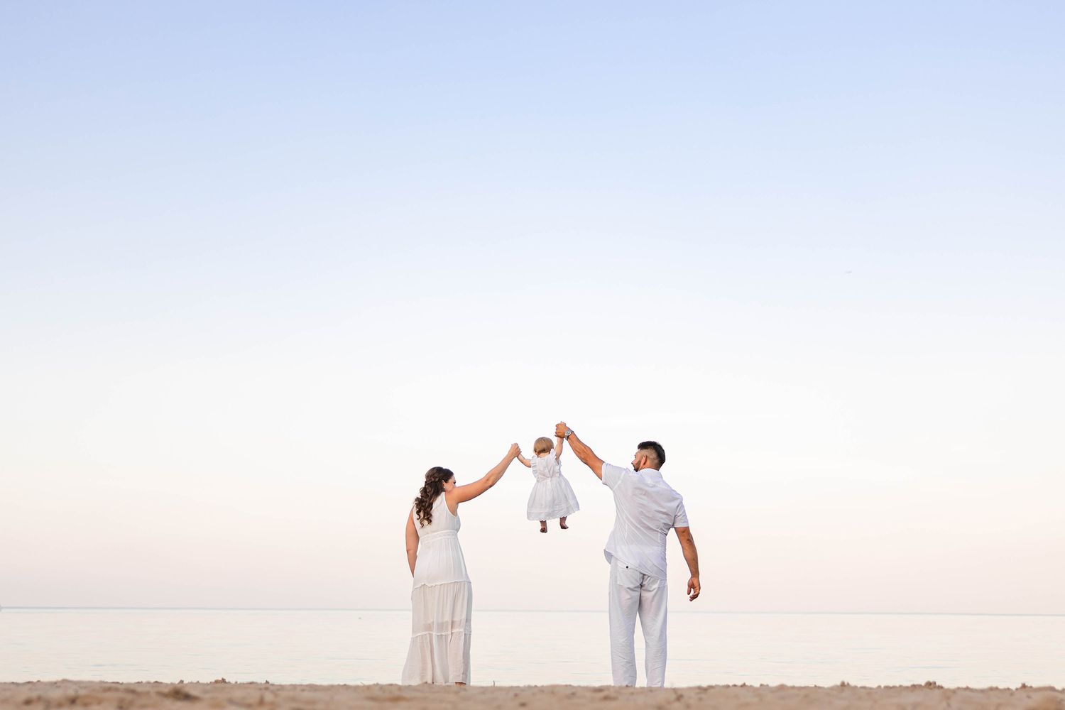 A couple in white outfits holds up a small child against a serene sky backdrop at sunset.