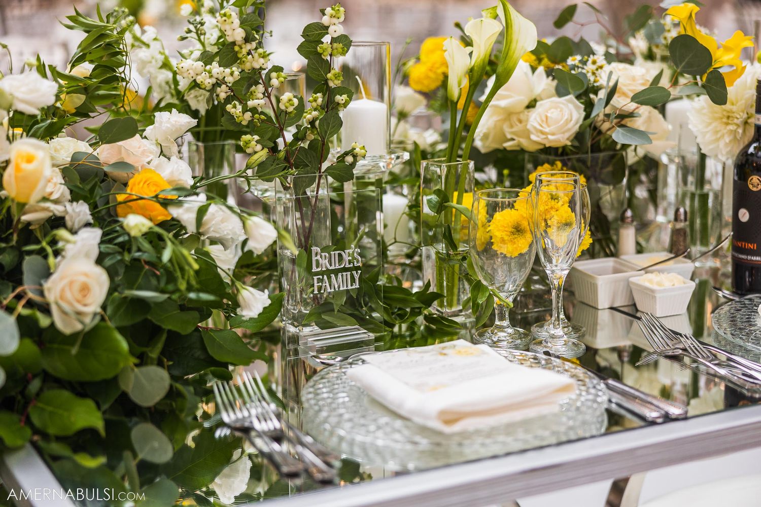 Wedding table setting with yellow and white roses in glass vases surrounded by elegant place settings.