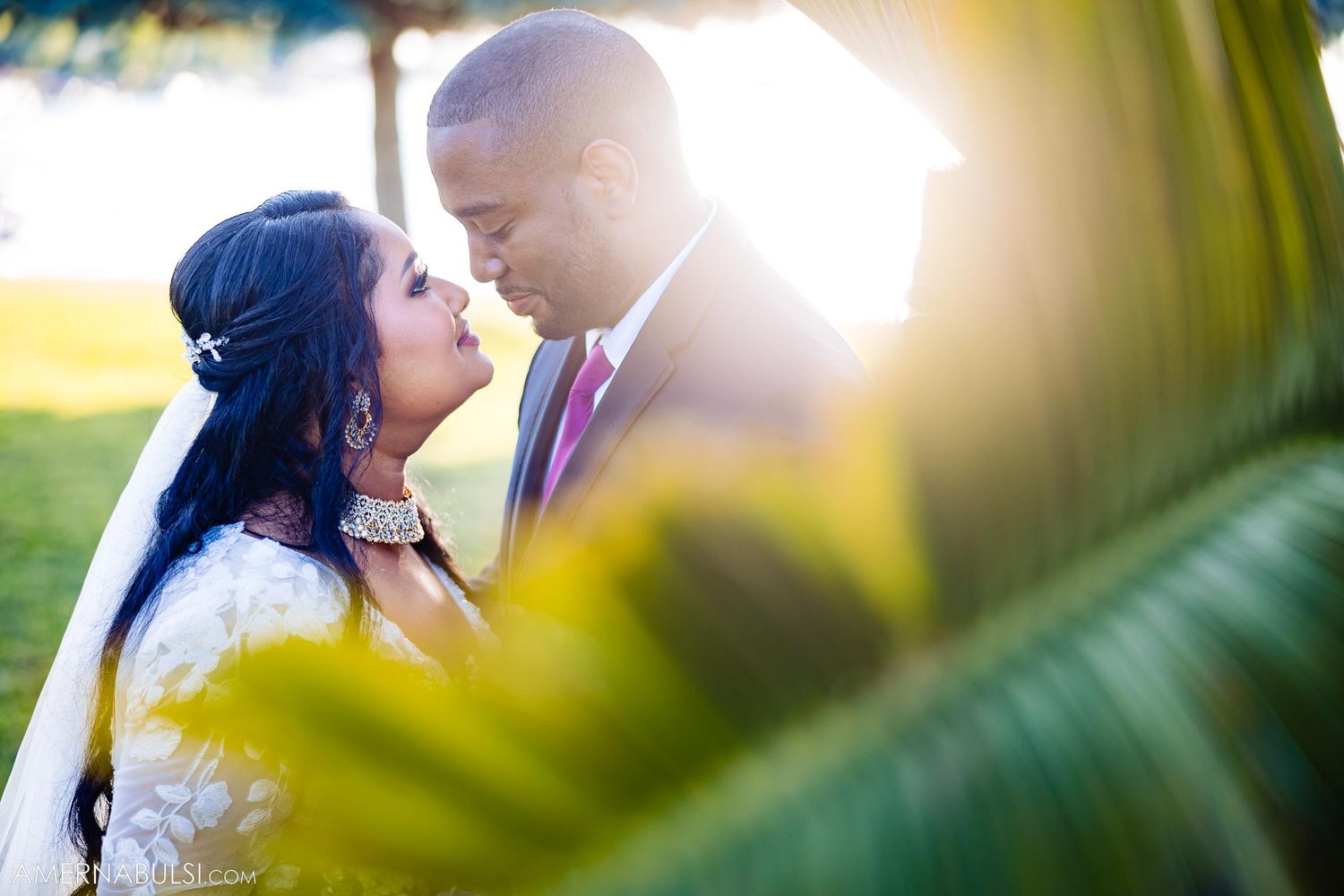 A romantic wedding portrait captured through palm fronds with sunlight creating a warm glow.