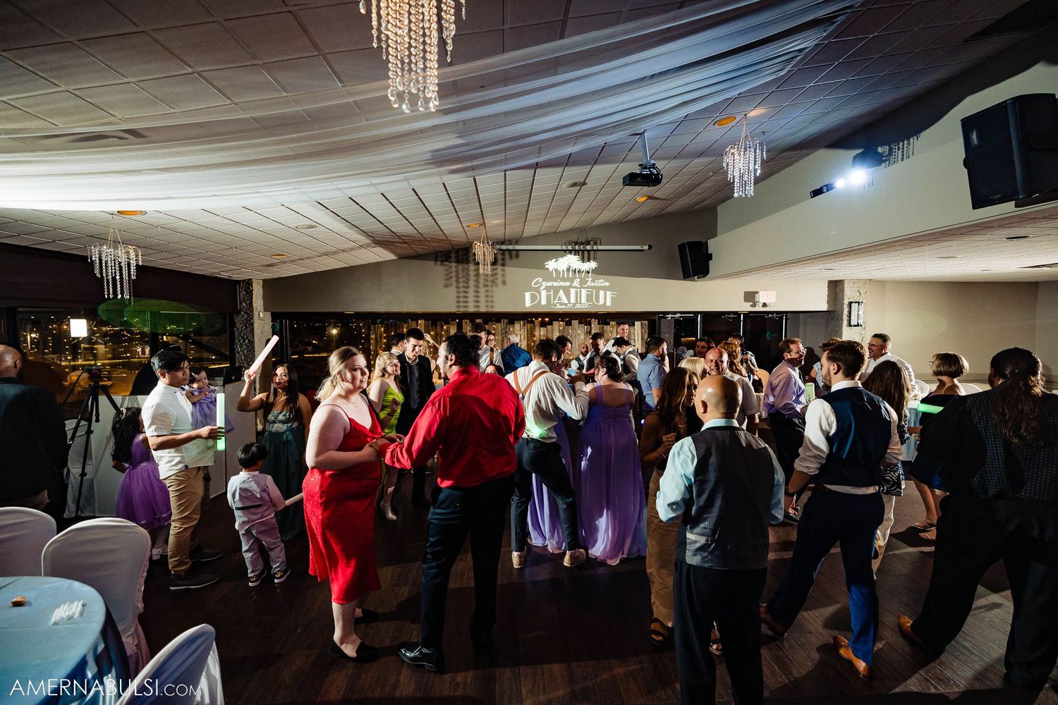 Large crowd dancing together in an event hall with crystal chandeliers and festive lighting.