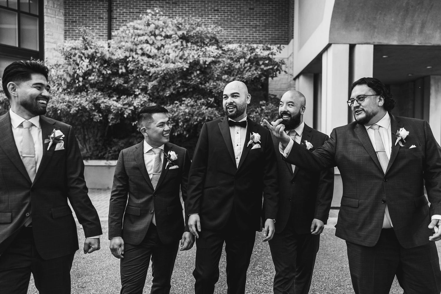 Groomsmen in black tuxedos share a laugh together outdoors in black and white photo.