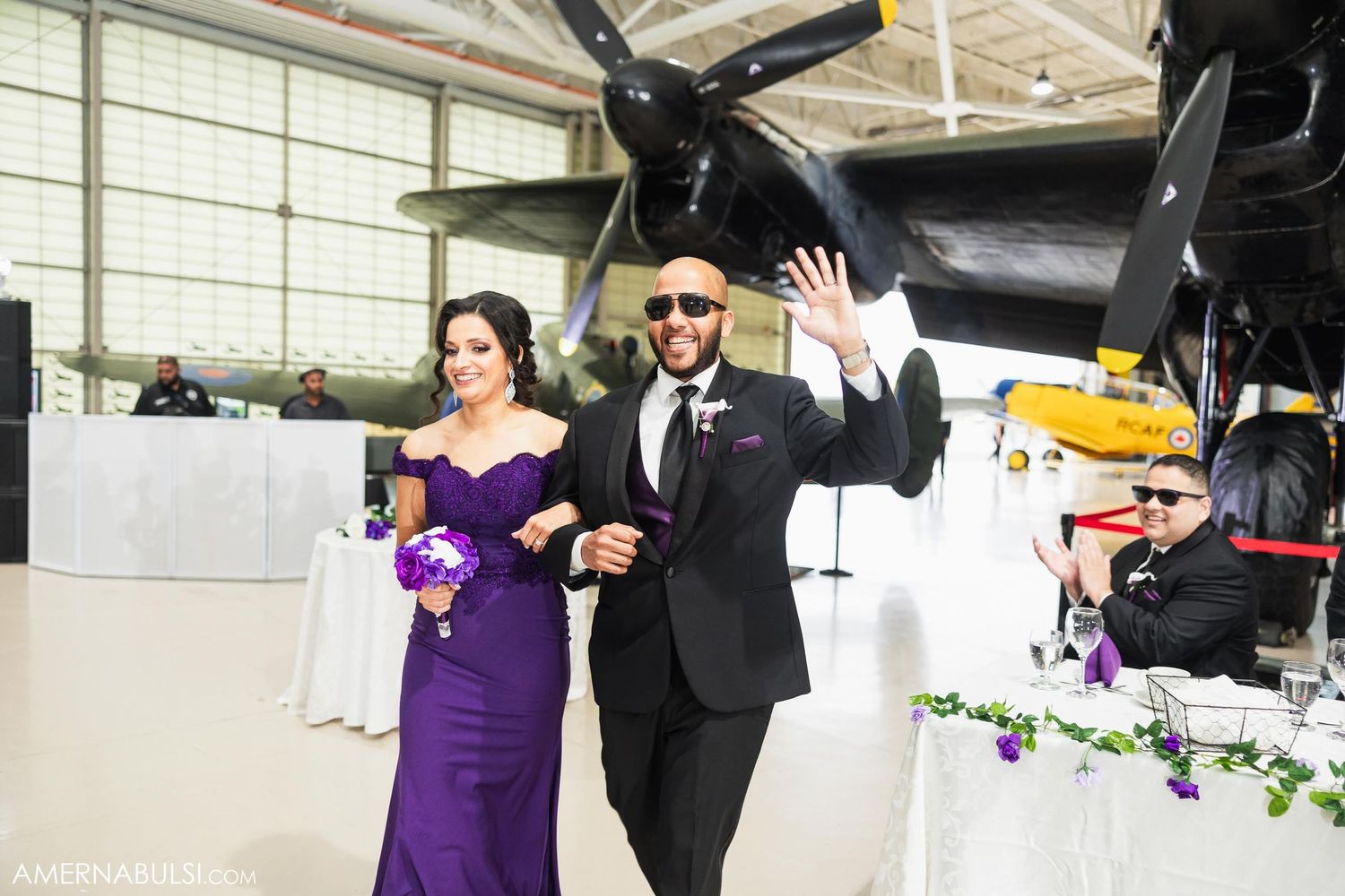 Wedding party members in purple and black attire walk through aircraft hangar.