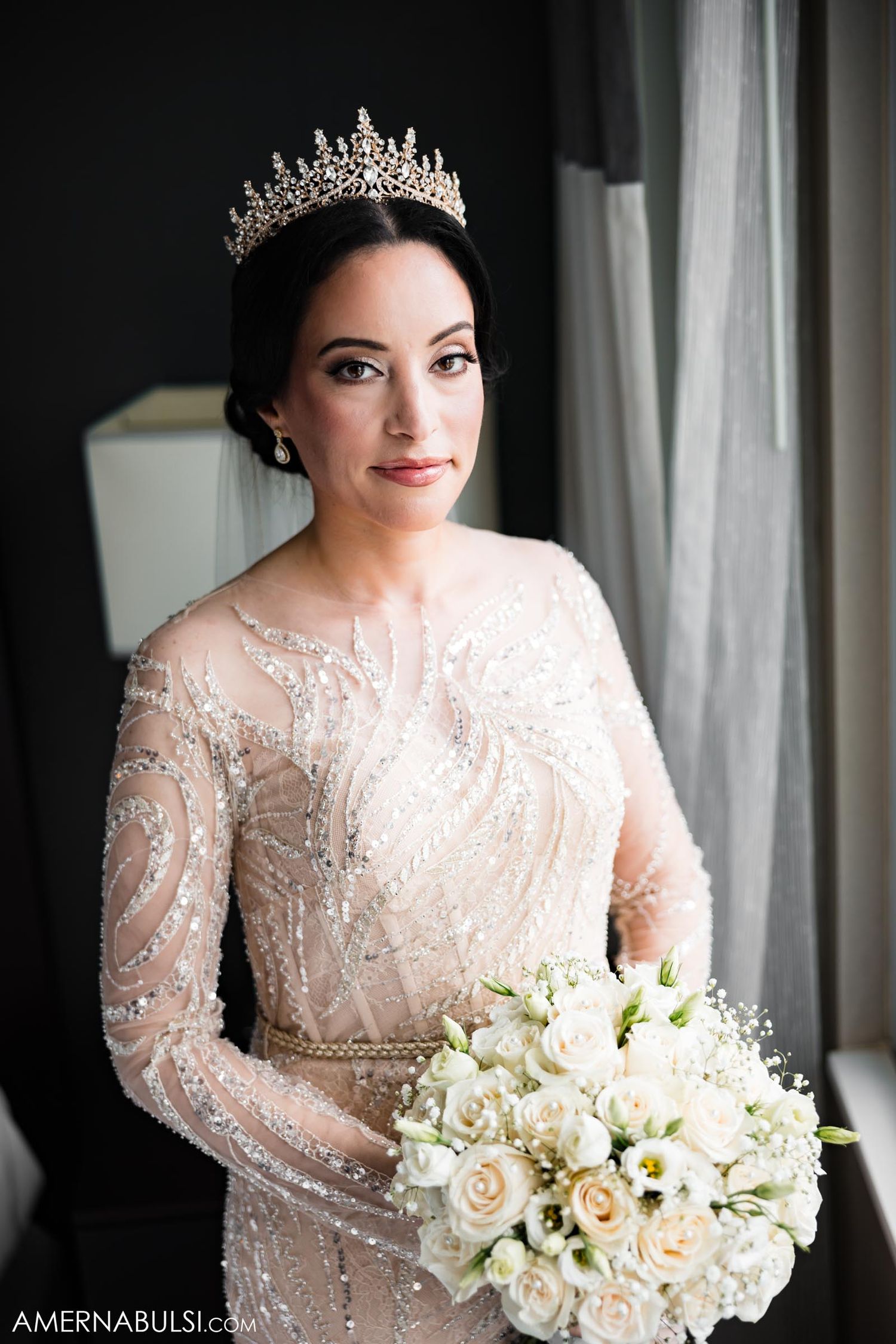 Bride wearing a crystal tiara and beaded gown holds white rose bouquet near window.