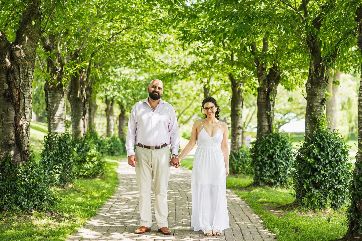 A couple in light summer wedding attire walks along a tree-lined path in a lush garden setting.